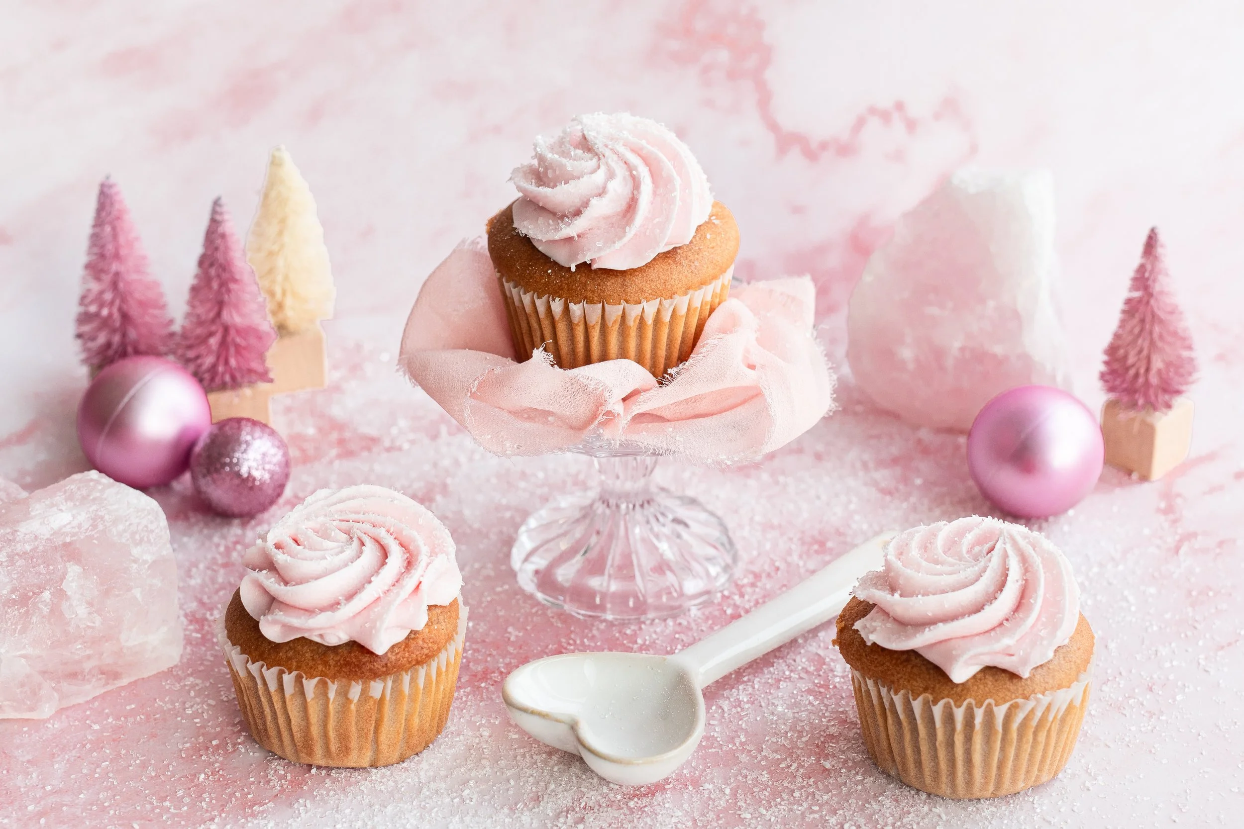 Pink frosted cupcakes on a glass stand surrounded by pink Christmas ornaments, mini trees, and white decorative items on a pink surface with a pink background.