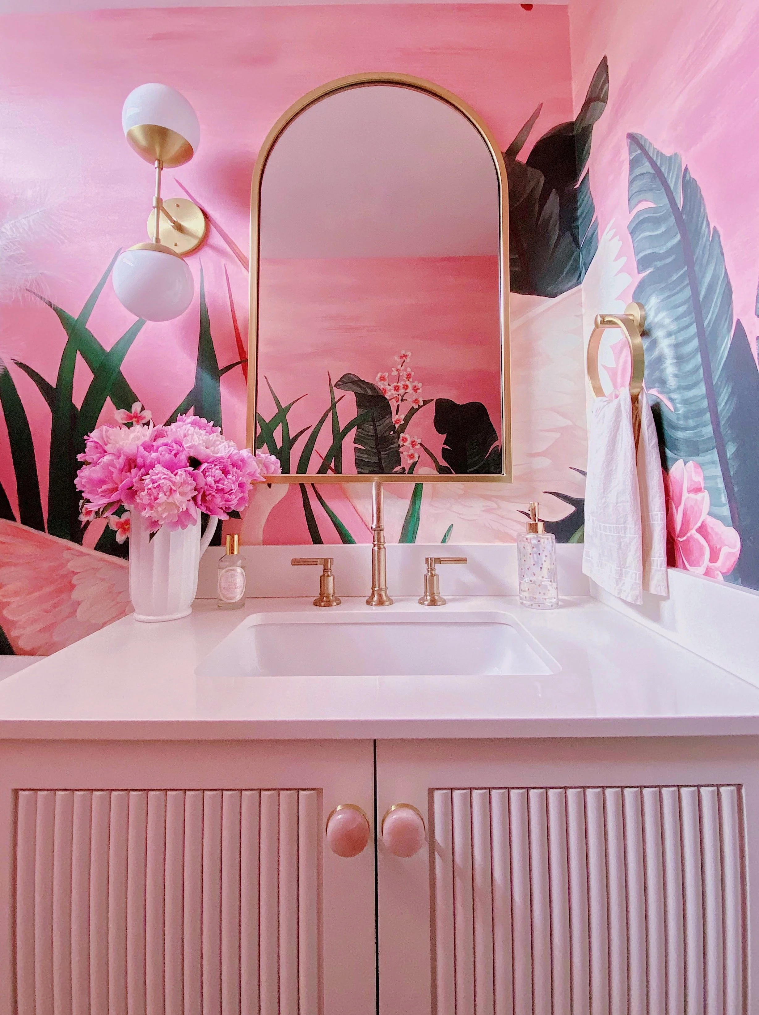 A bathroom vanity with a white sink, a gold faucet, and a cabinet with vertical pink ridges. On the countertop, there is a vase with pink flowers, a small decorative bottle, and a clear glass container. Above the sink, a curved mirror is mounted on a