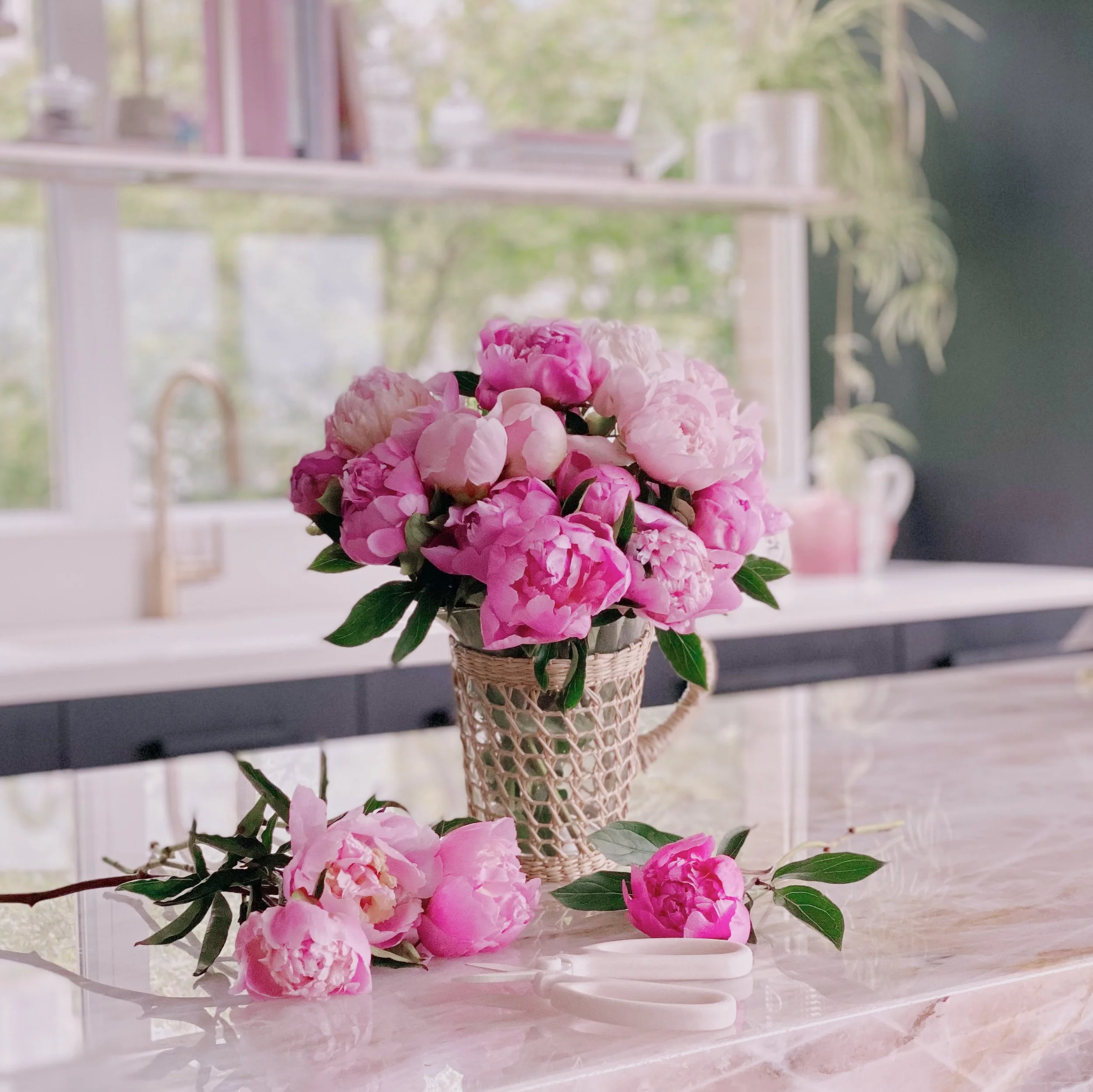 Pink peonies in a wicker basket on a marble countertop, with a window in the background showing trees.