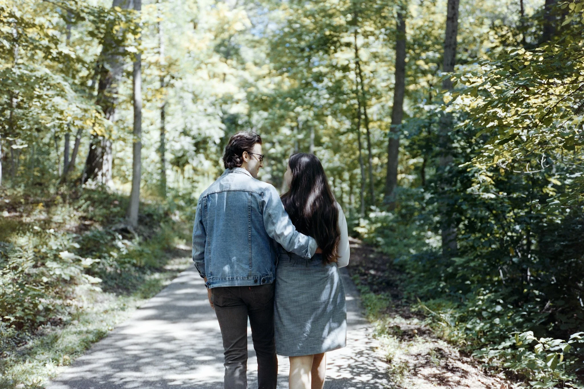 A man and woman walking together in a wooded park, holding arms and enjoying a sunny day.