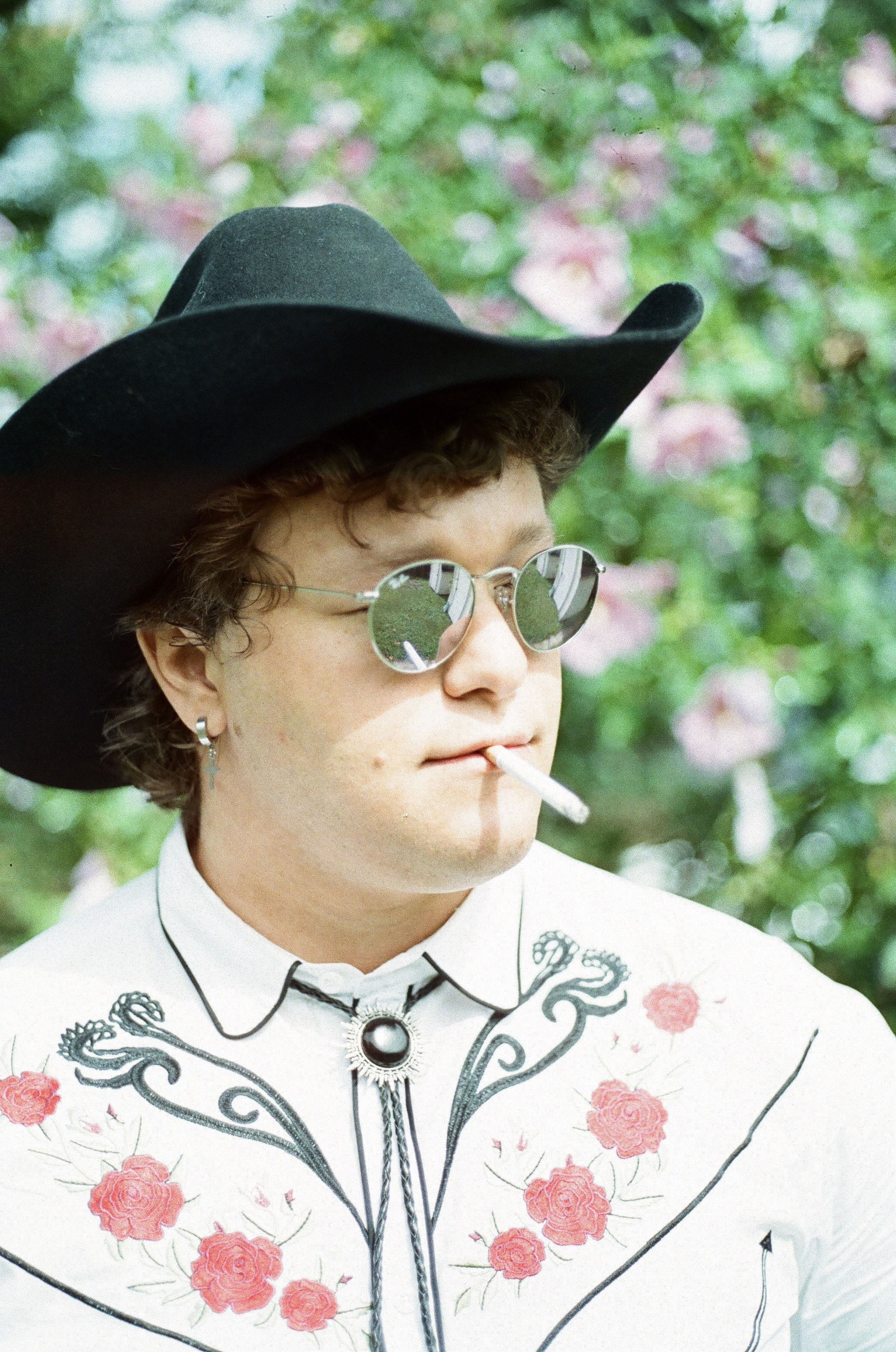A young man with curly brown hair, wearing mirrored sunglasses, a black wide-brimmed hat, and a white embroidered shirt with red roses, is smoking a cigarette outdoors with a blurred green and pink floral background.