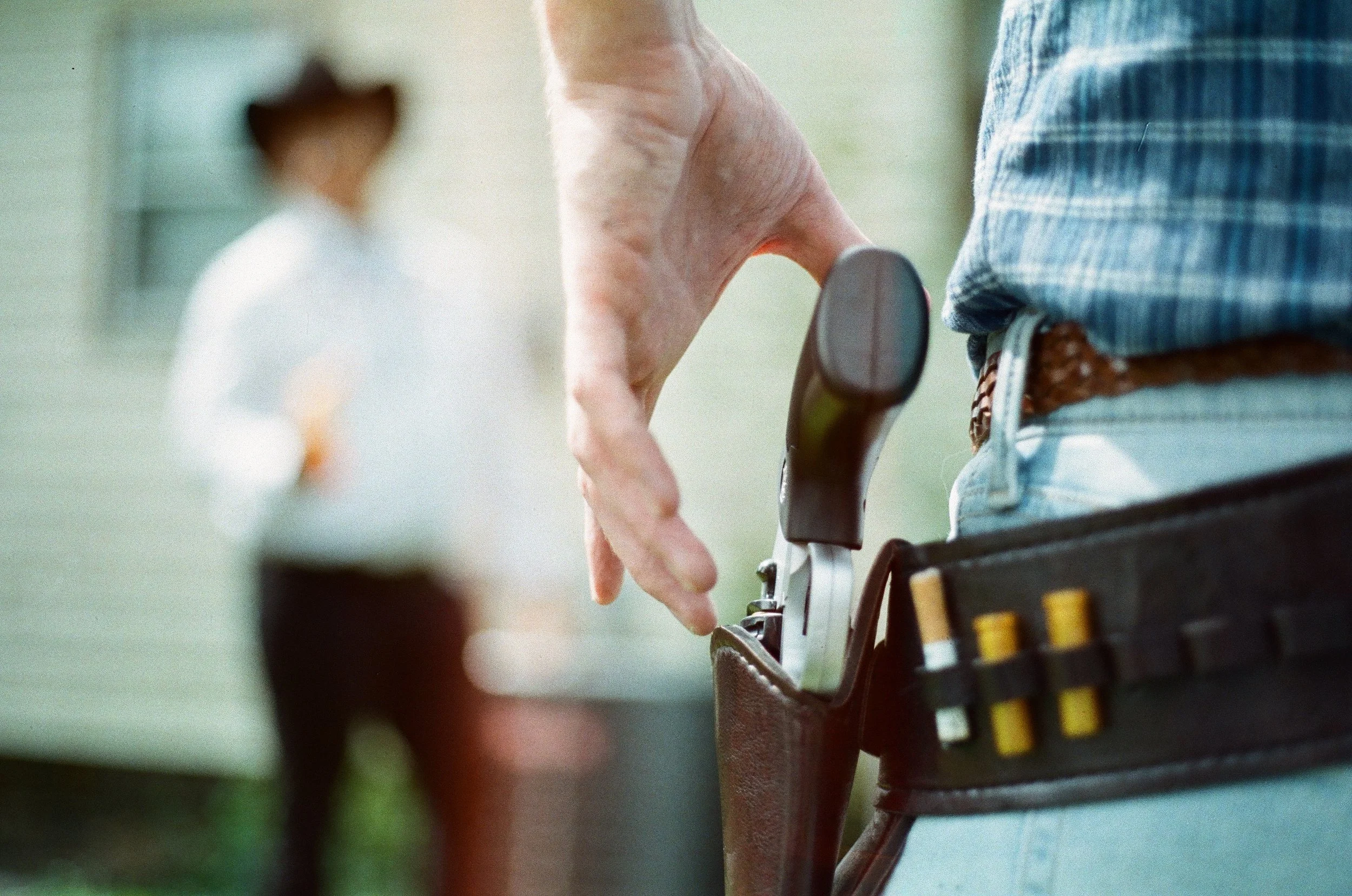 Close-up of a person wearing a blue plaid shirt, placing a pistol into a black holster on their waist belt, with a blurred figure of a woman in the background.