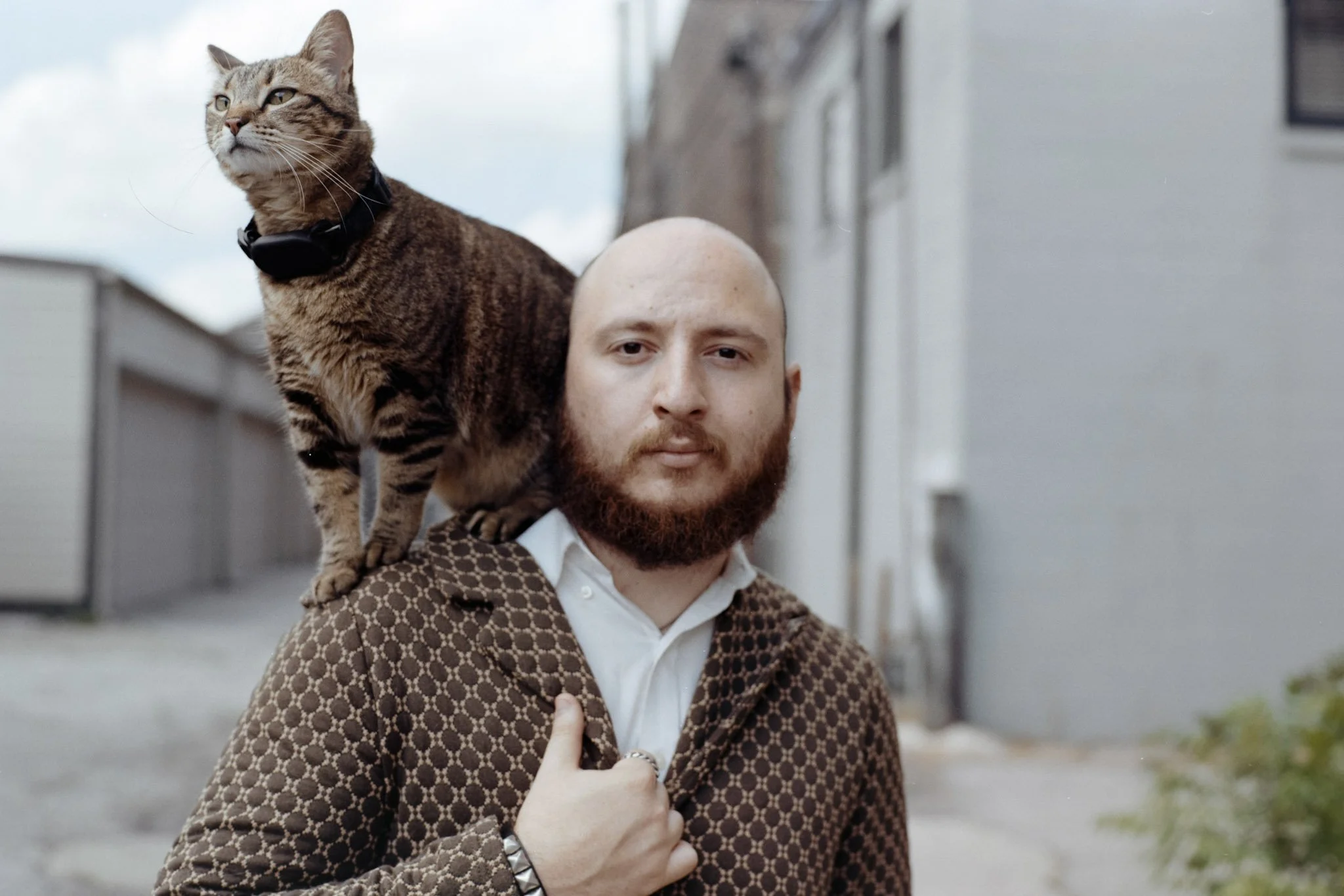A man with a fedora-like hat carrying a tabby cat on his shoulder in an alleyway.