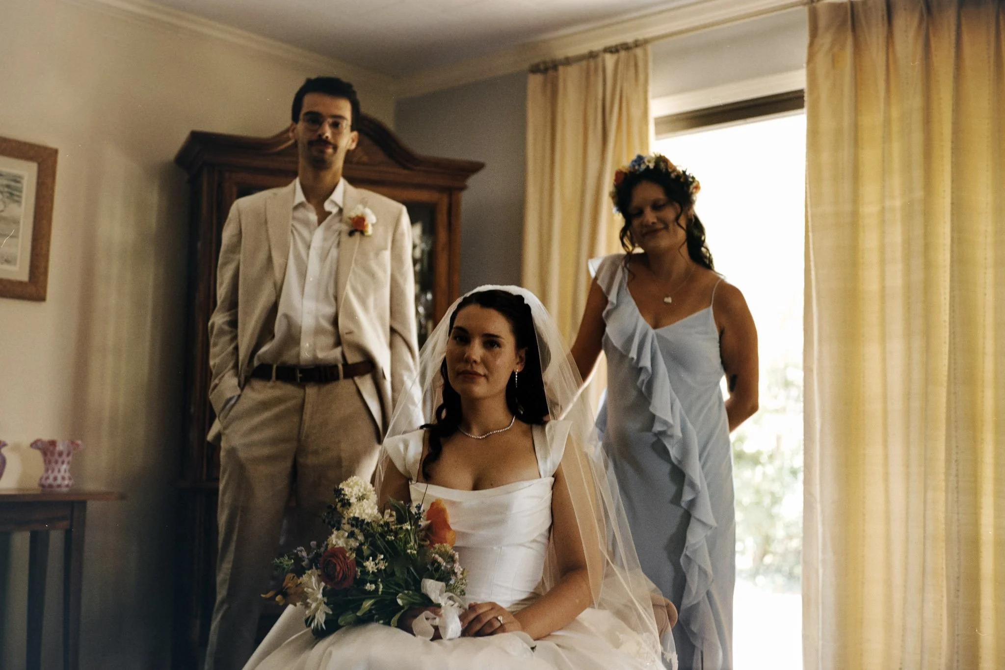 A bride in a white wedding dress sitting with a bouquet of flowers, flanked by a man and a woman, both dressed formally, inside a room with yellow curtains and sunlight coming through the window.