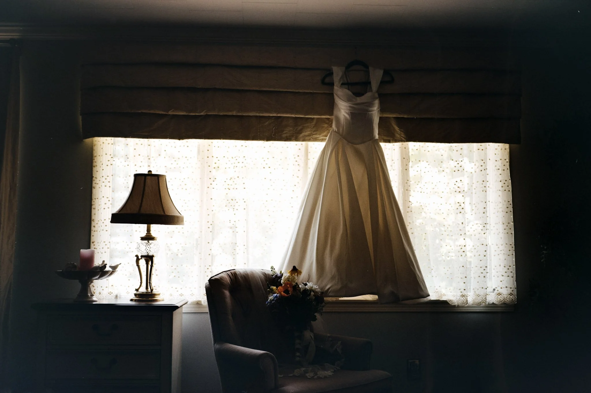 A wedding dress hangs on a window in a dimly lit room, with a table lamp, flowers, and chairs in the foreground.