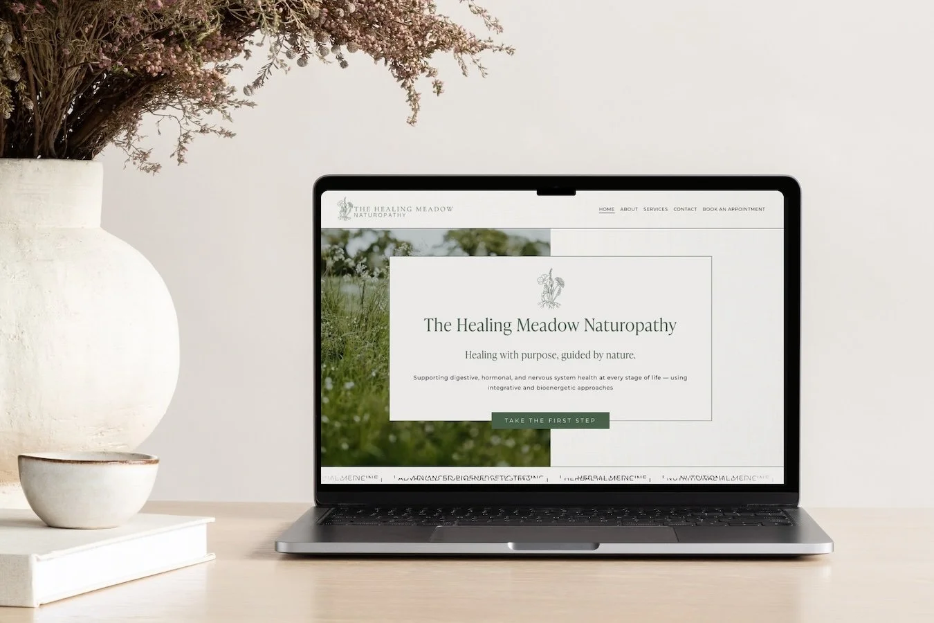 A laptop displays a website for The Healing Meadow Naturopathy on a desk, with a white vase with pink flowers and a smaller white bowl on the left.