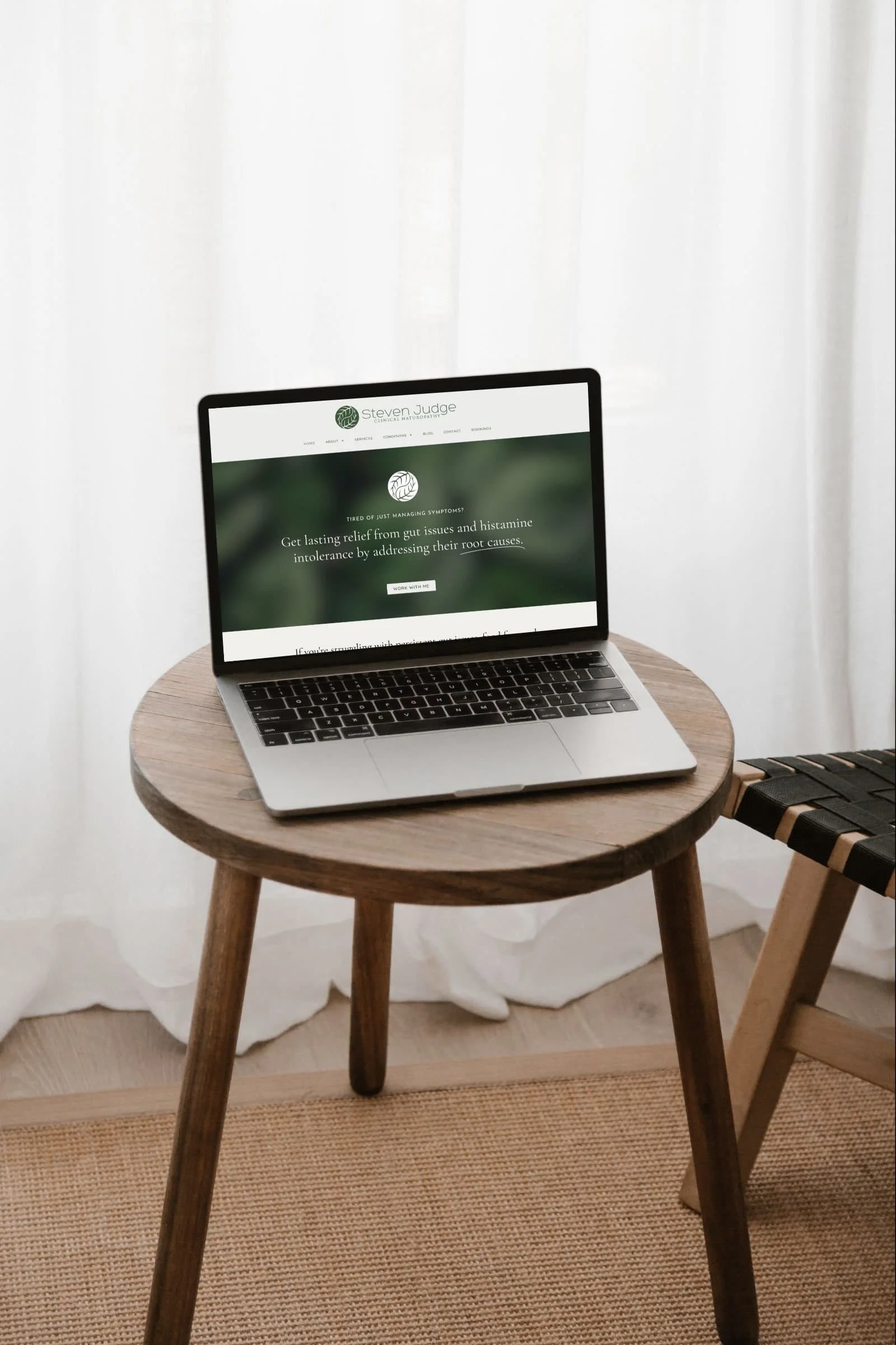 A wooden round table with a laptop open to Steven Judge Naturopath website about gut health, placed in a room with white curtains and a small woven rug underneath.