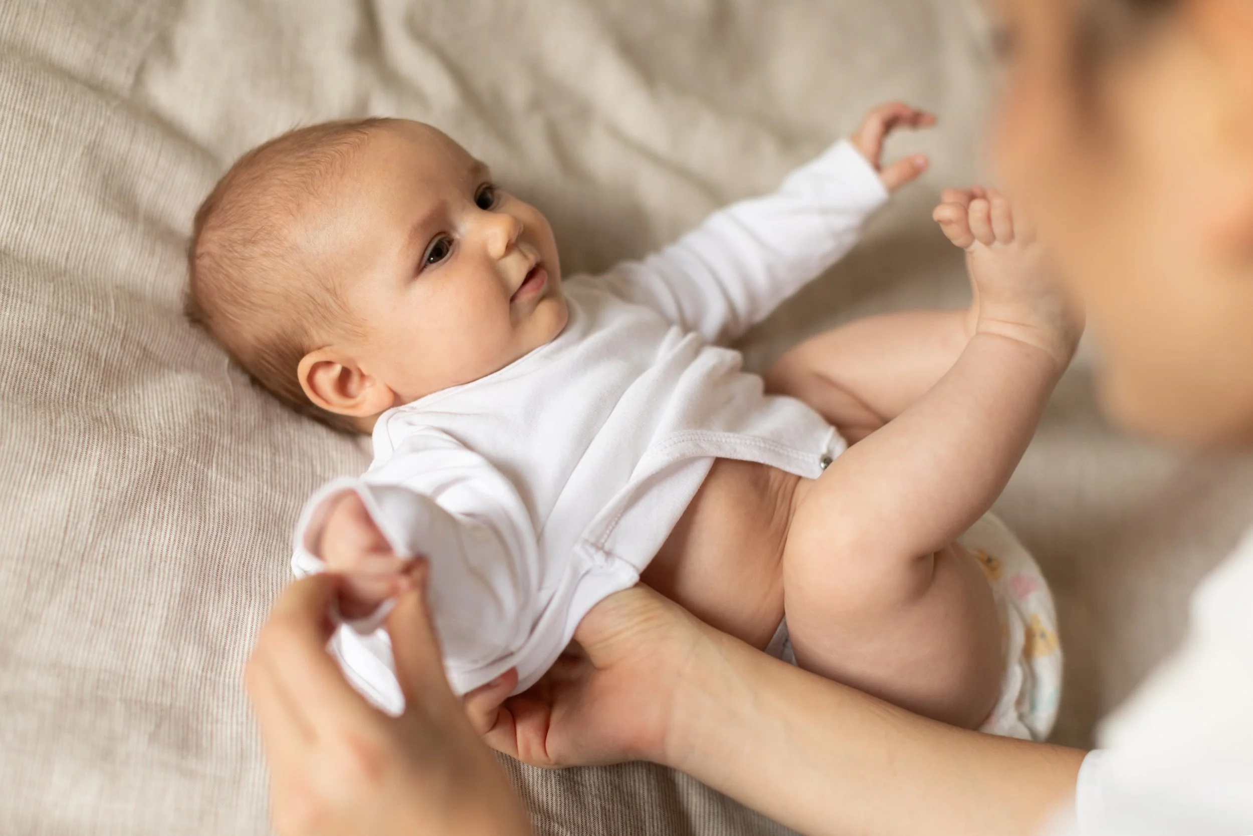 A baby lying on a beige surface, looking up at a person holding her. The baby is wearing a white shirt and diaper, with arms stretched out.