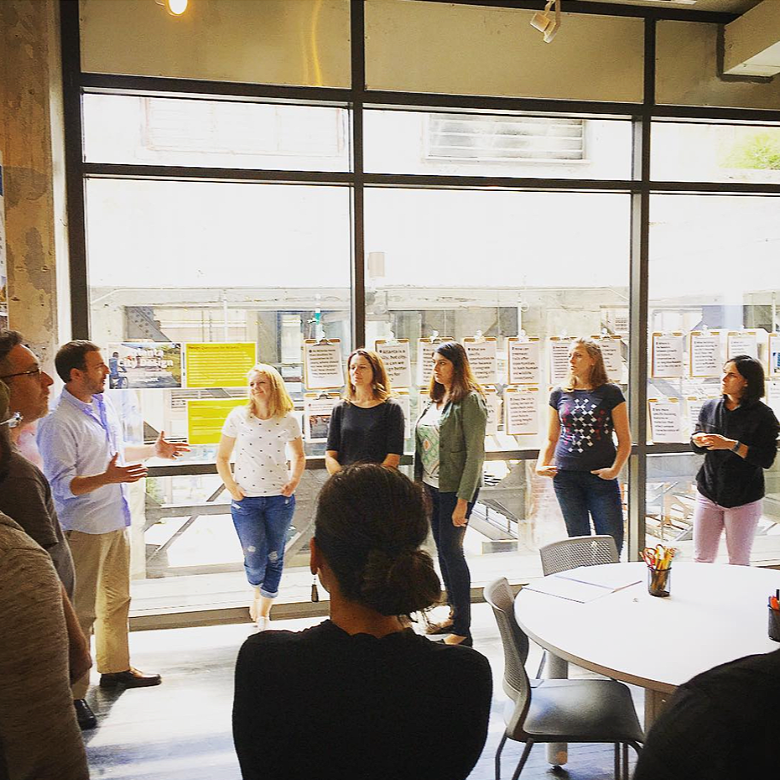 People standing indoors near large windows with posters on display in the background, some engaging in conversation, others listening.