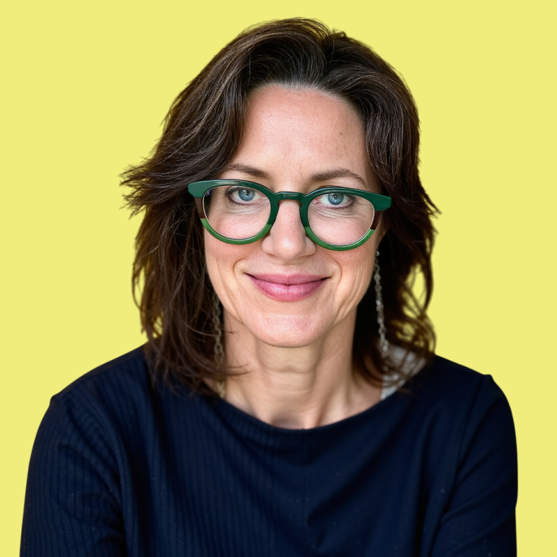 A woman with brown, shoulder-length hair, wearing green glasses and earrings, smiling, against a yellow background.