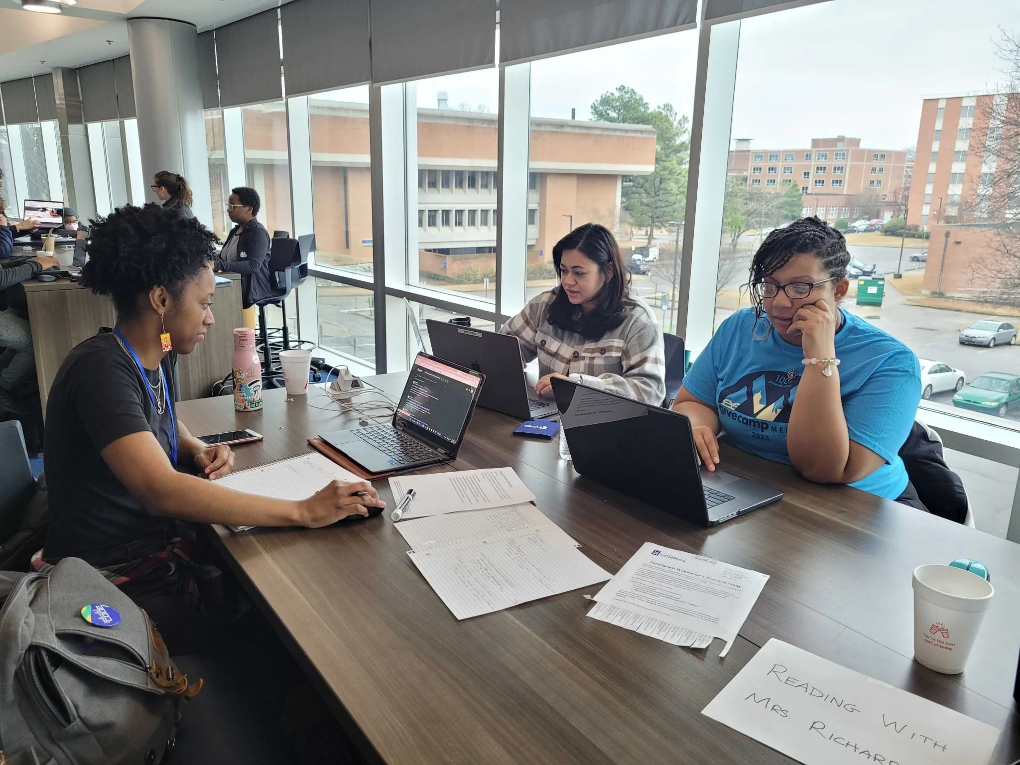 Three women sitting at a table in a library or study area, working on laptops and studying with papers and notebooks in front of them. The table has a sign that reads 'Reading with Mrs. Richard.' Large windows behind them show a parking lot and trees outside.