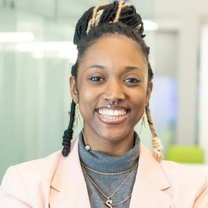A young Black woman with dreadlocks and a septum piercing, smiling and looking at the camera, wearing a light pink blazer over a gray turtleneck, with layered necklaces, in a modern office setting.