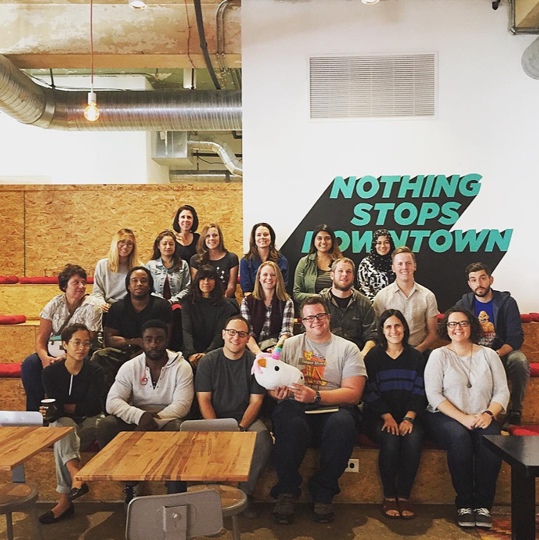 Group of people posing for a photo in a modern, industrial-style space with a mix of wood and open ceiling design. They are seated and standing on tiered wooden steps, with a sign on the wall that reads "Nothing Stops Downtown."