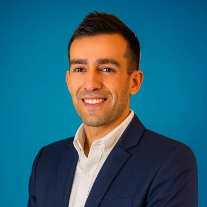 A man wearing a dark suit and white shirt smiling against a blue background.