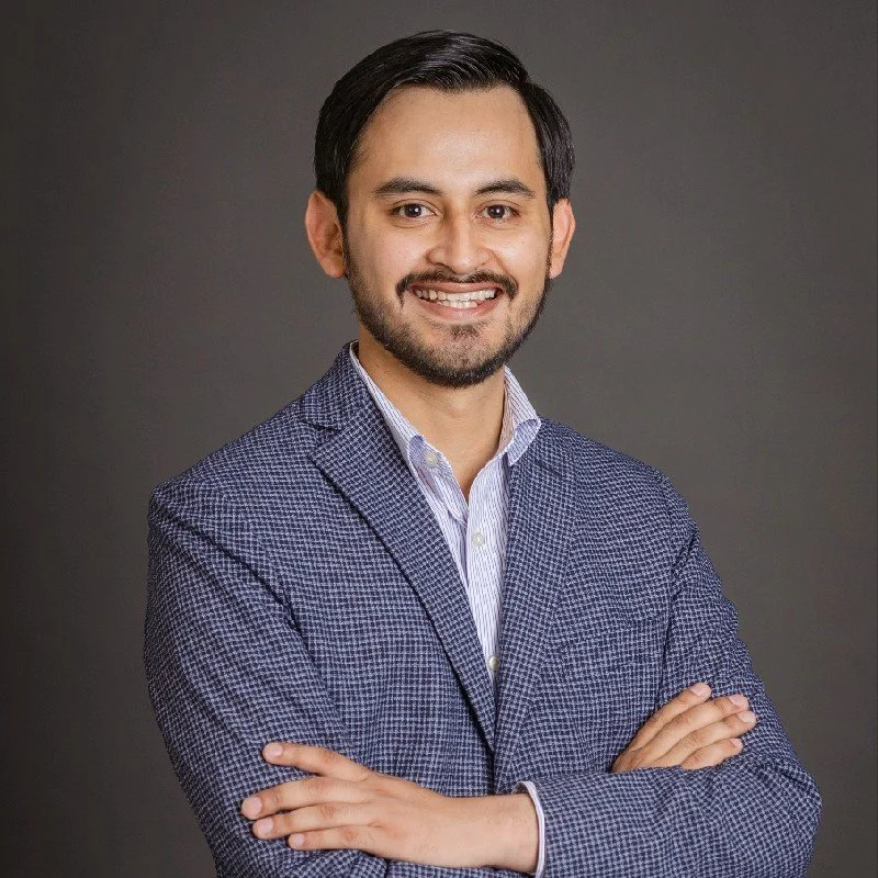 A man with dark hair and a beard wearing a blue patterned blazer and a light-colored collared shirt, standing with arms crossed against a gray background and smiling.