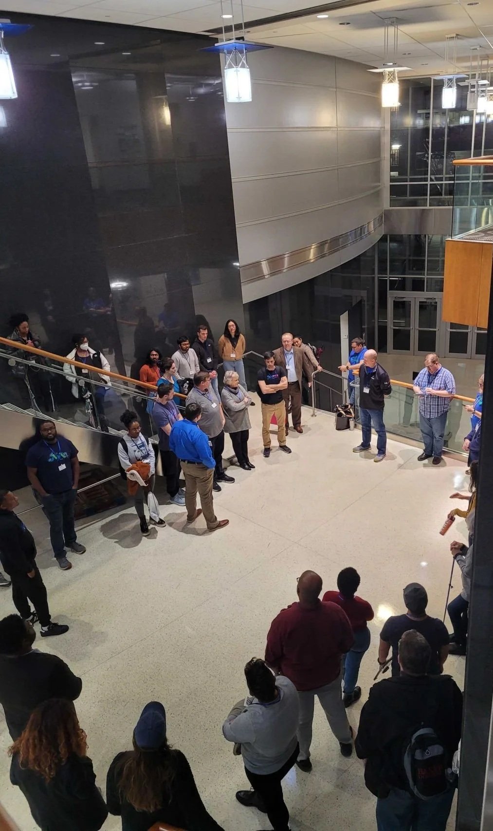 A group of people gathered in a modern building lobby, standing in a semicircle, listening to a speaker in the center.