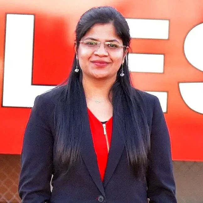 A woman with long black hair, glasses, earrings, wearing a dark blazer over a red top, standing in front of a red wall with partial white letters.