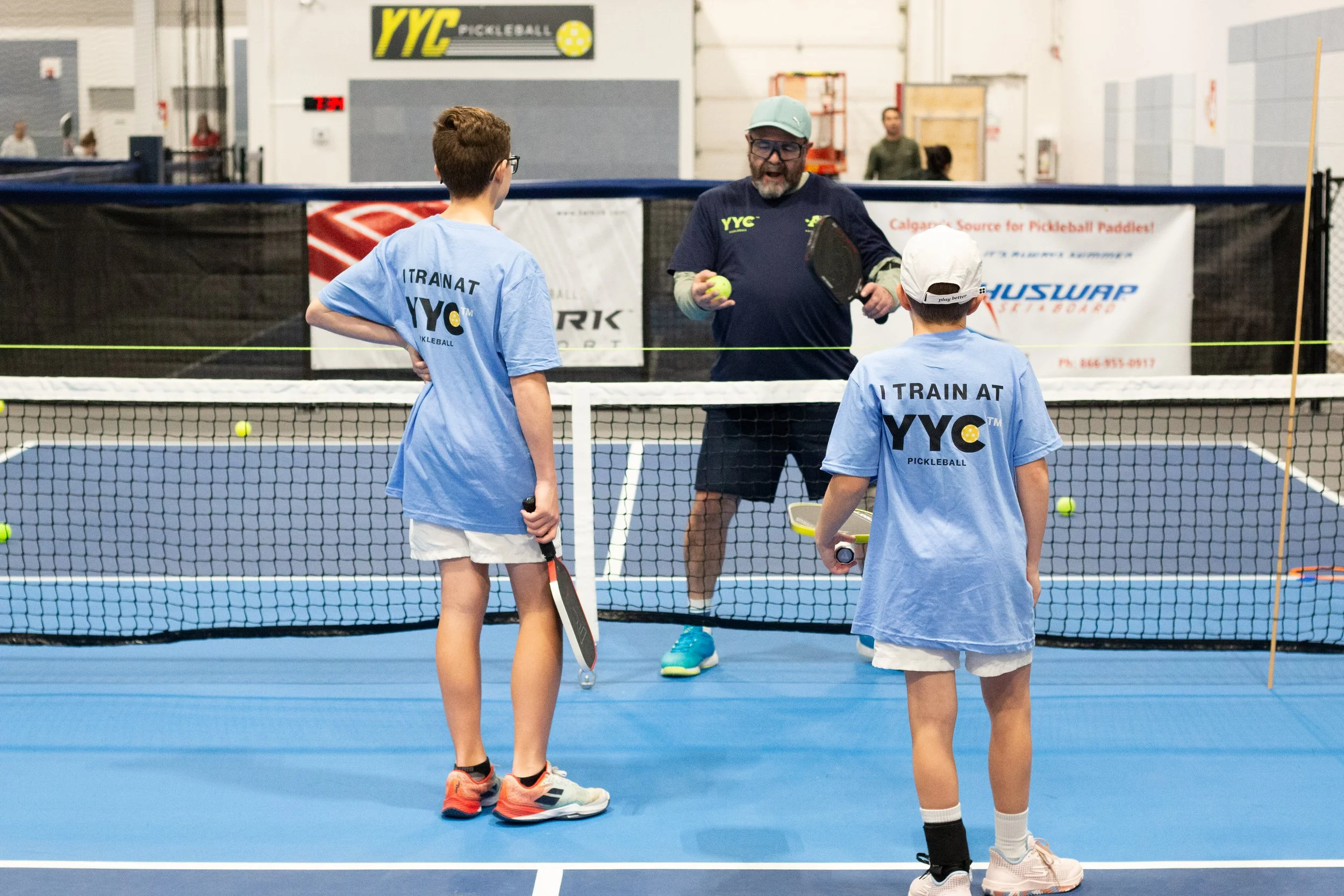 Two young children with paddle tennis rackets talking to a coach on an indoor paddleball court, wearing blue T-shirts that say 'I TRAIN AT YYC'.