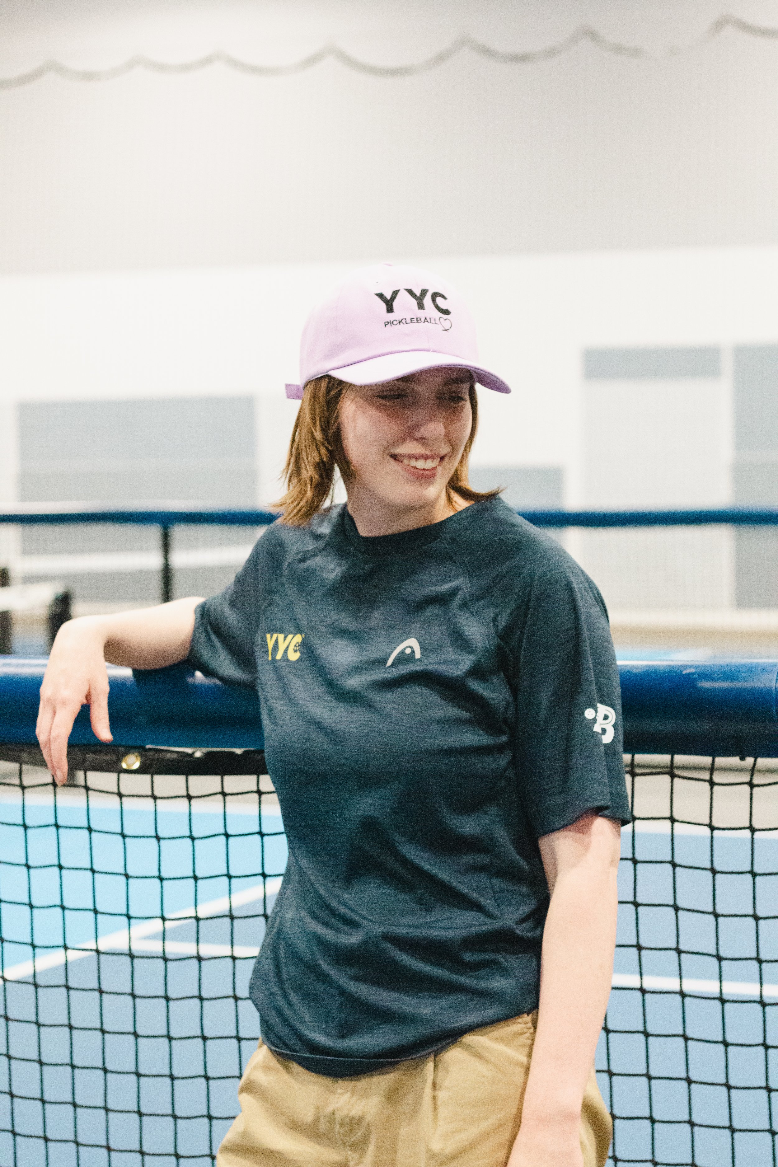 A young woman standing near a tennis net inside an indoor sports facility, wearing a pink hat with 'YYC Pickleball' and a dark T-shirt with the same logo, smiling and looking to her right.