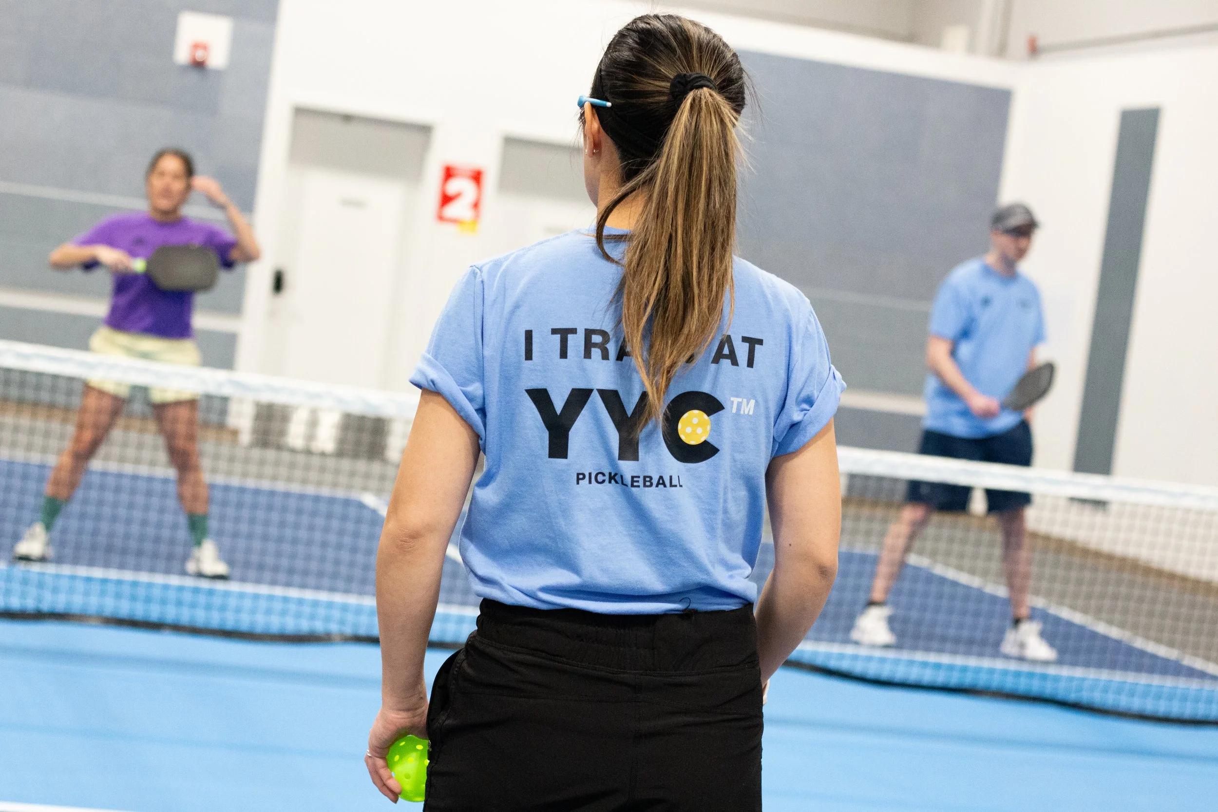 A woman in a blue shirt watching a pickleball game with two players on the court, one woman in purple and one man in blue, playing at an indoor sports facility.