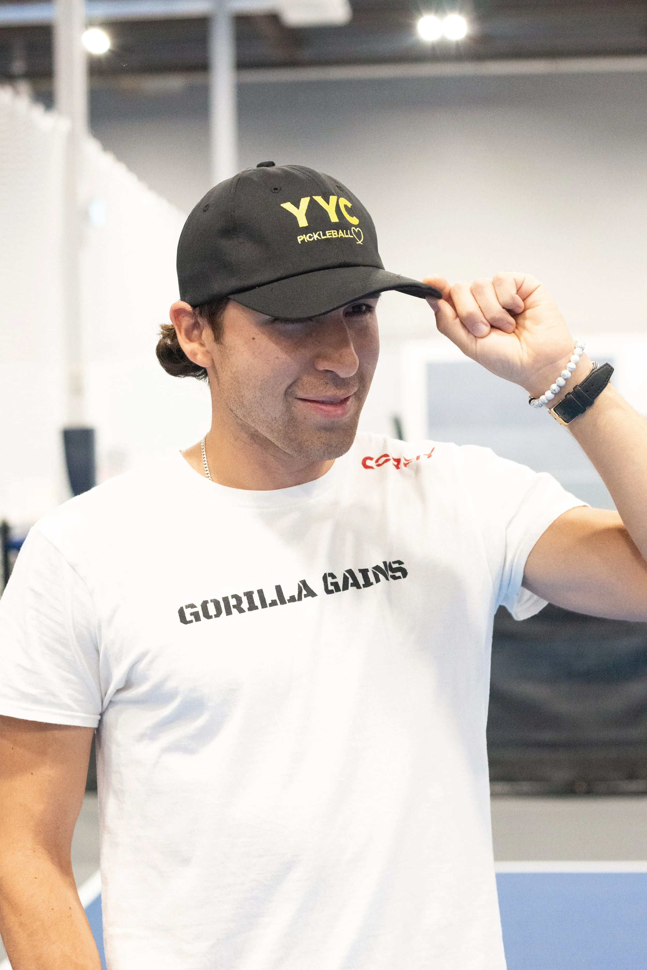 A man wearing a black cap with yellow letters, a white T-shirt with 'Gorilla Gains' printed on it, and a beaded bracelet, adjusting his cap inside a fitness or sports facility.