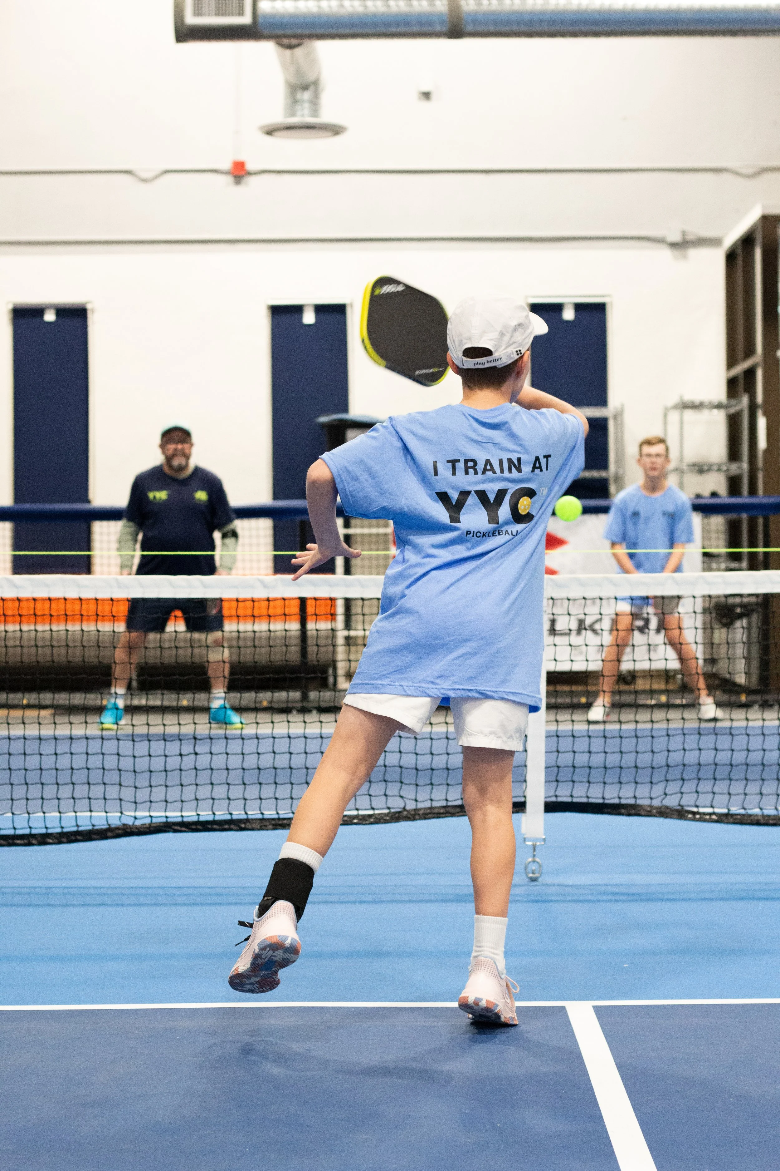 Child playing pickleball on indoor court with a boy in the background and an adult at the far end of the court.