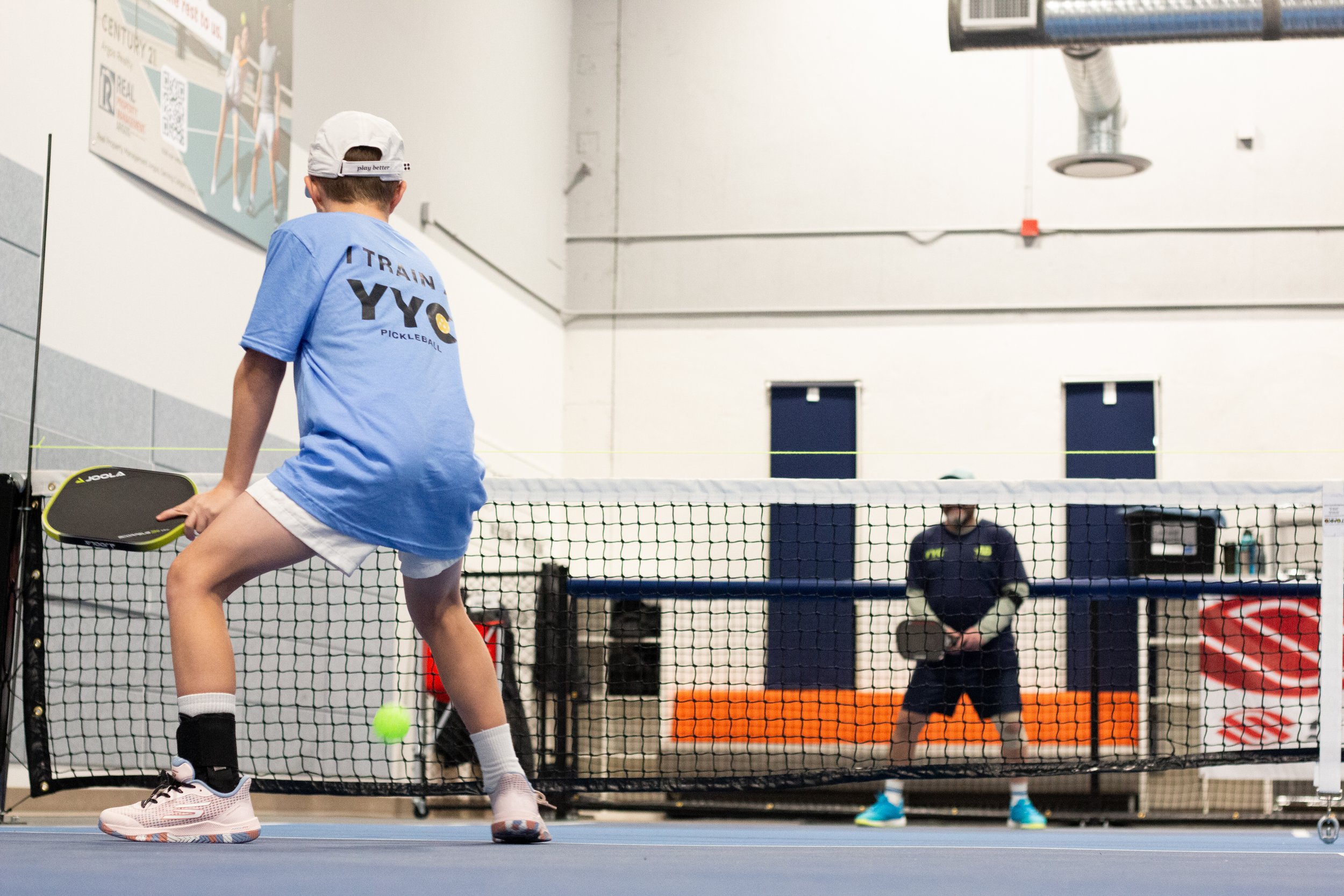 Young boy playing pickleball indoors, about to hit a tennis ball with a paddle, with a net, and a man standing in the background.