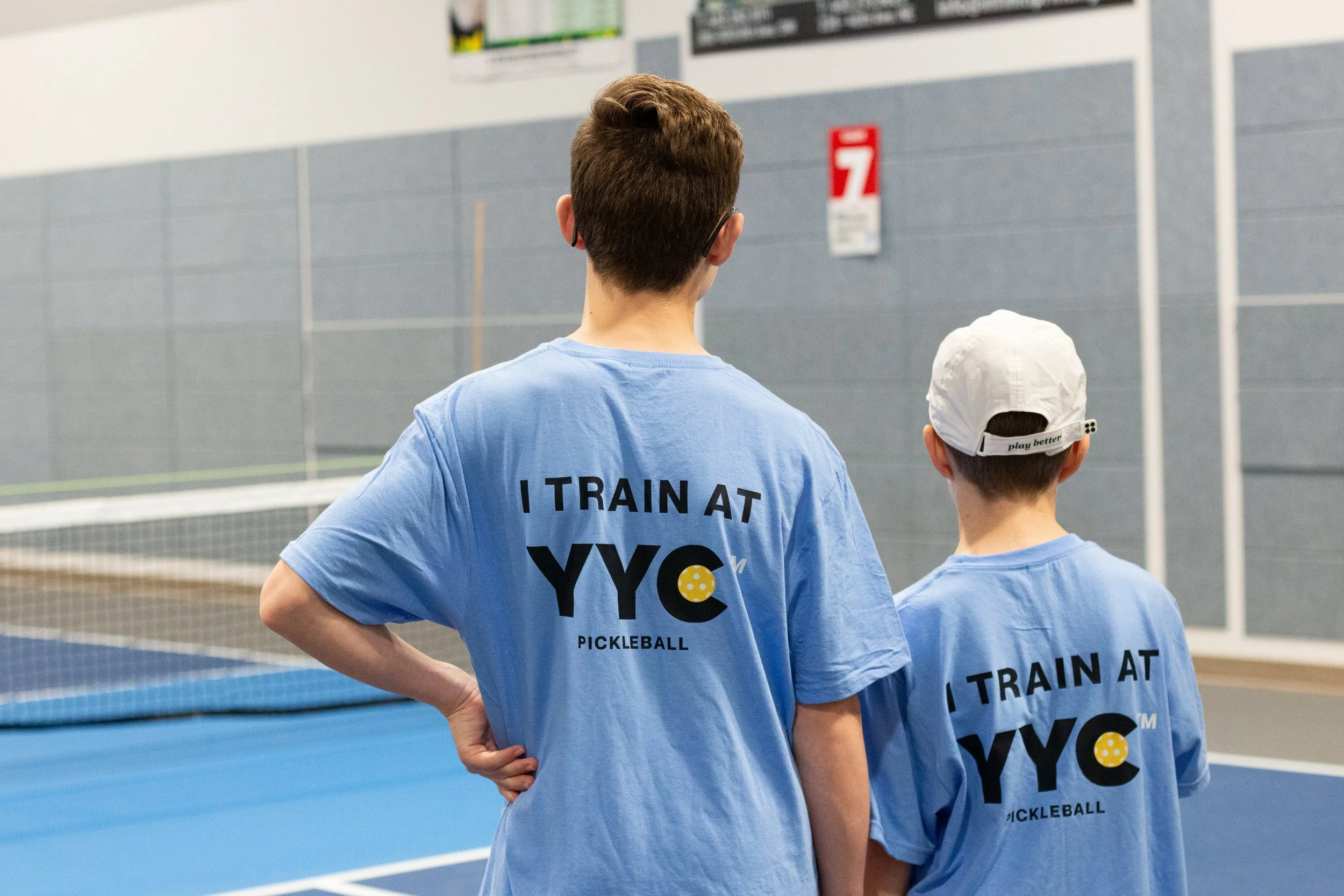 Two young boys in blue t-shirts with the text "I TRAIN AT YYC PICKLEBALL" printed on the back, standing on an indoor pickleball court.