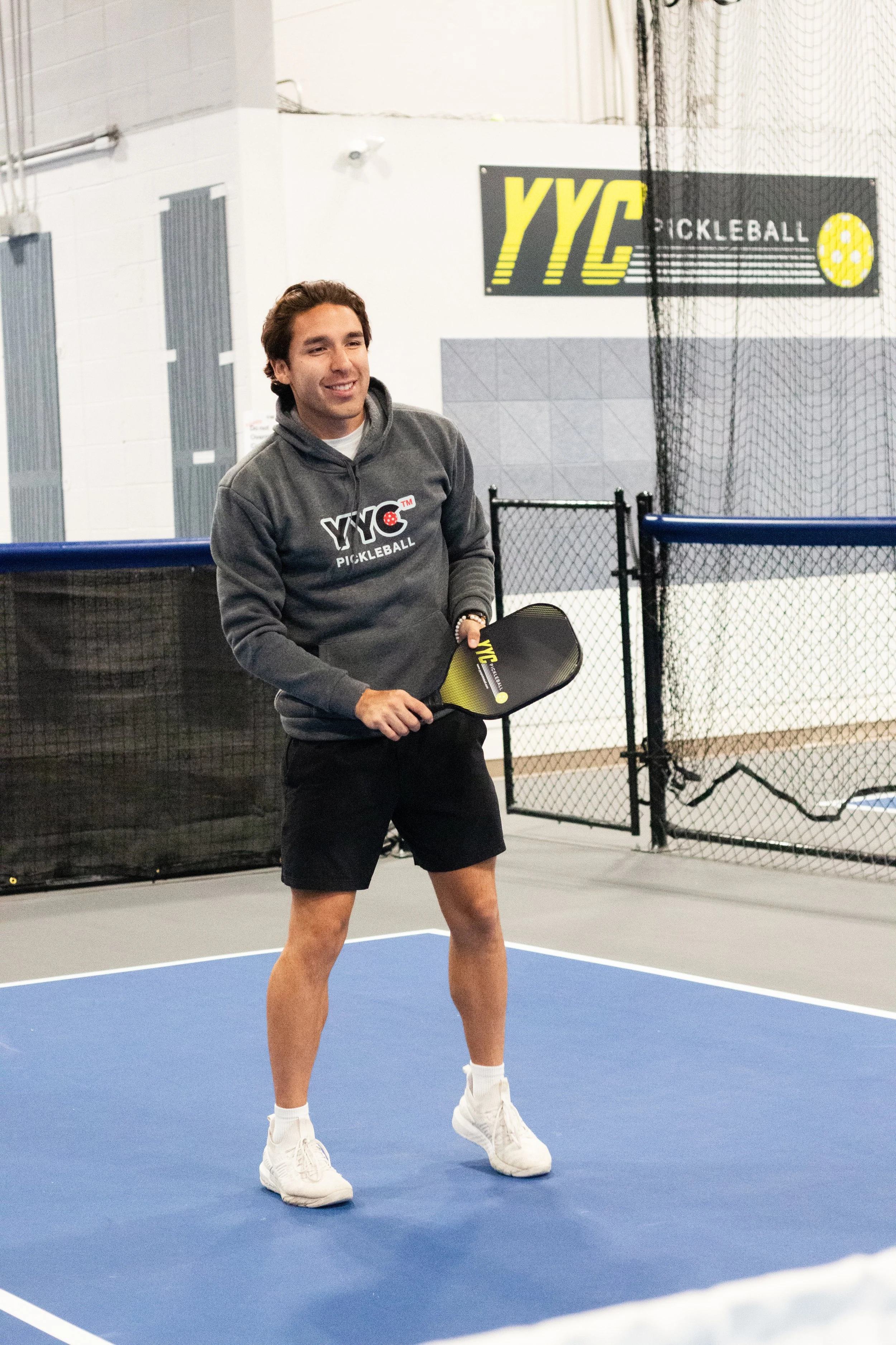 A man standing on a pickleball court holding a pickleball paddle, wearing a gray hoodie with 'YVC Pickleball' logo, black shorts, and white sneakers.