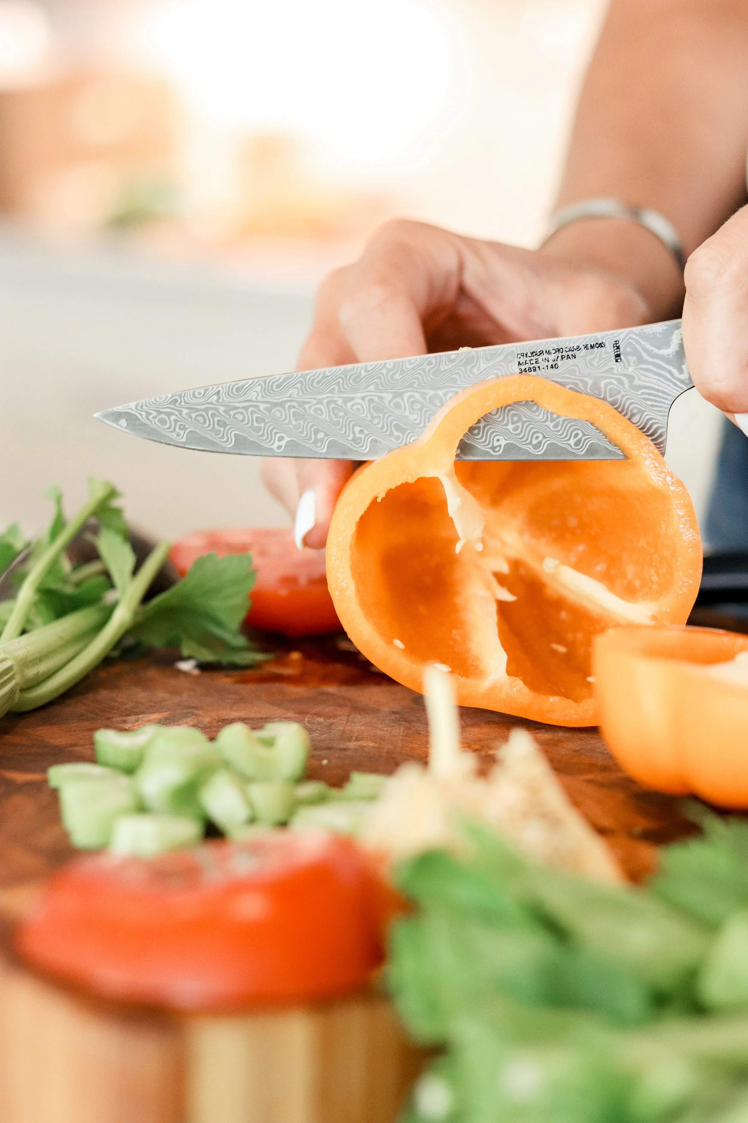 Close-up of a chef slicing a tomato with a patterned Japanese kitchen knife on a wooden cutting board, with celery and herbs nearby.