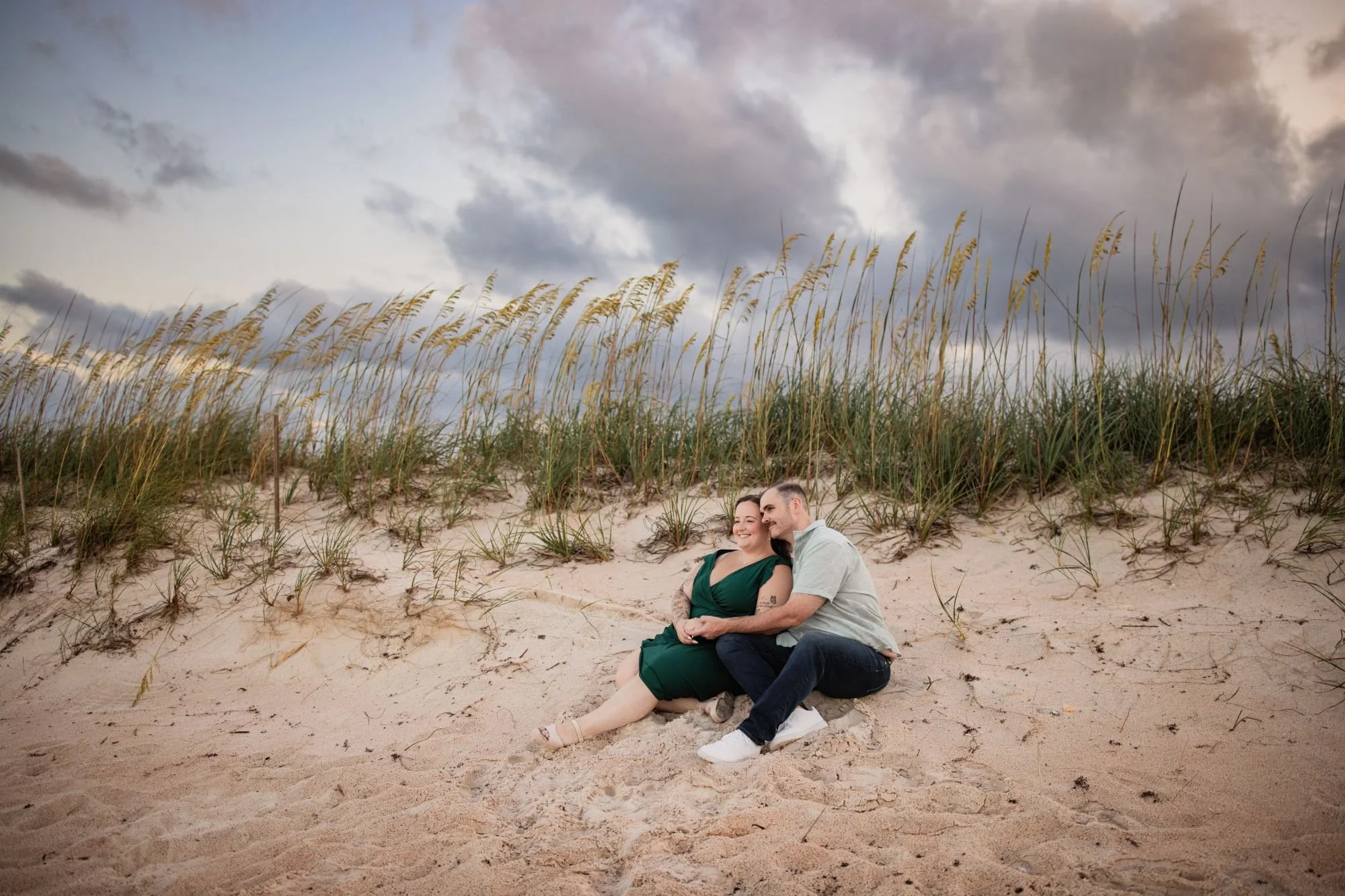 Engaged couple sitting together beneath sea oats and dramatic clouds at Washington Oaks beach.
