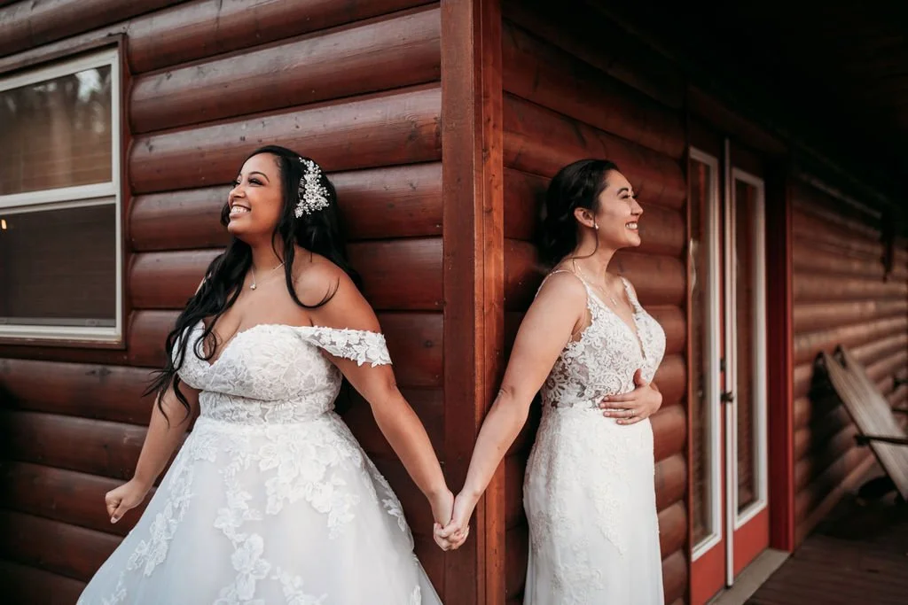 Two brides holding hands beside a rustic log cabin before their Blue Ridge Mountain wedding