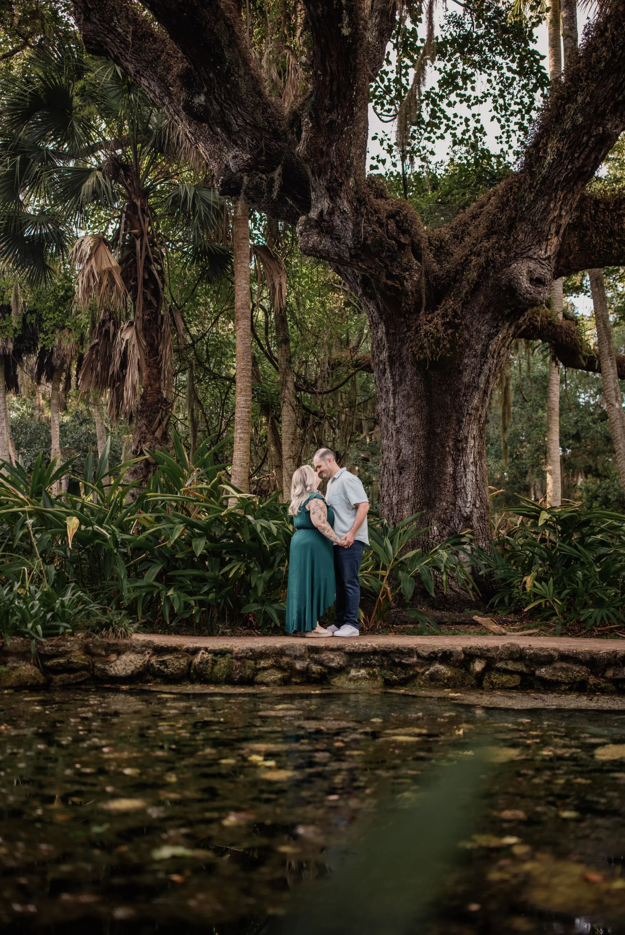 Couple standing beneath a massive oak tree beside a garden pond at Washington Oaks State Park.