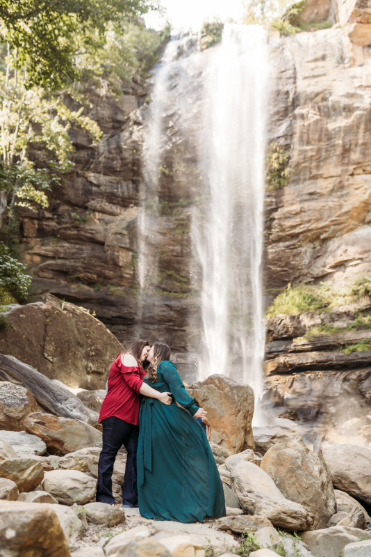 Two women kissing by a waterfall in red and emerald outfits