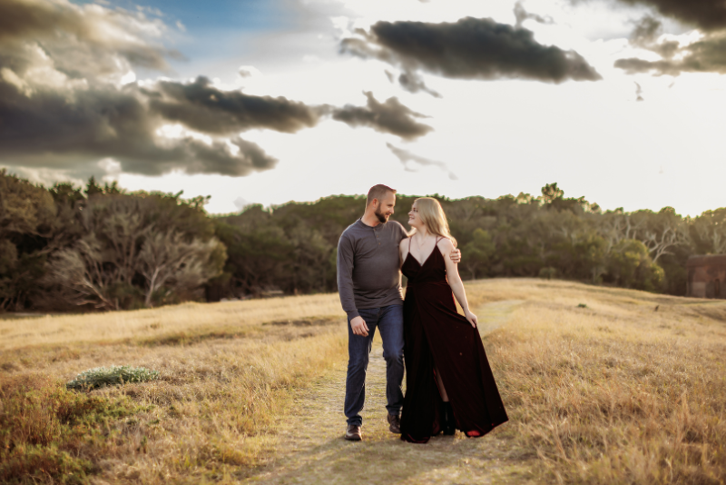Couple walking through a field in burgundy and charcoal outfits beneath a dramatic sky