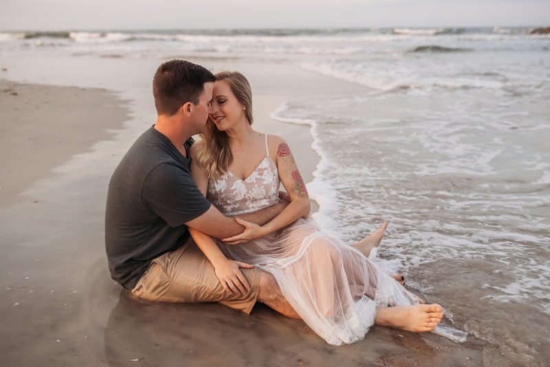 Couple sitting at the shoreline in soft neutral outfits during a relaxed beach photo session