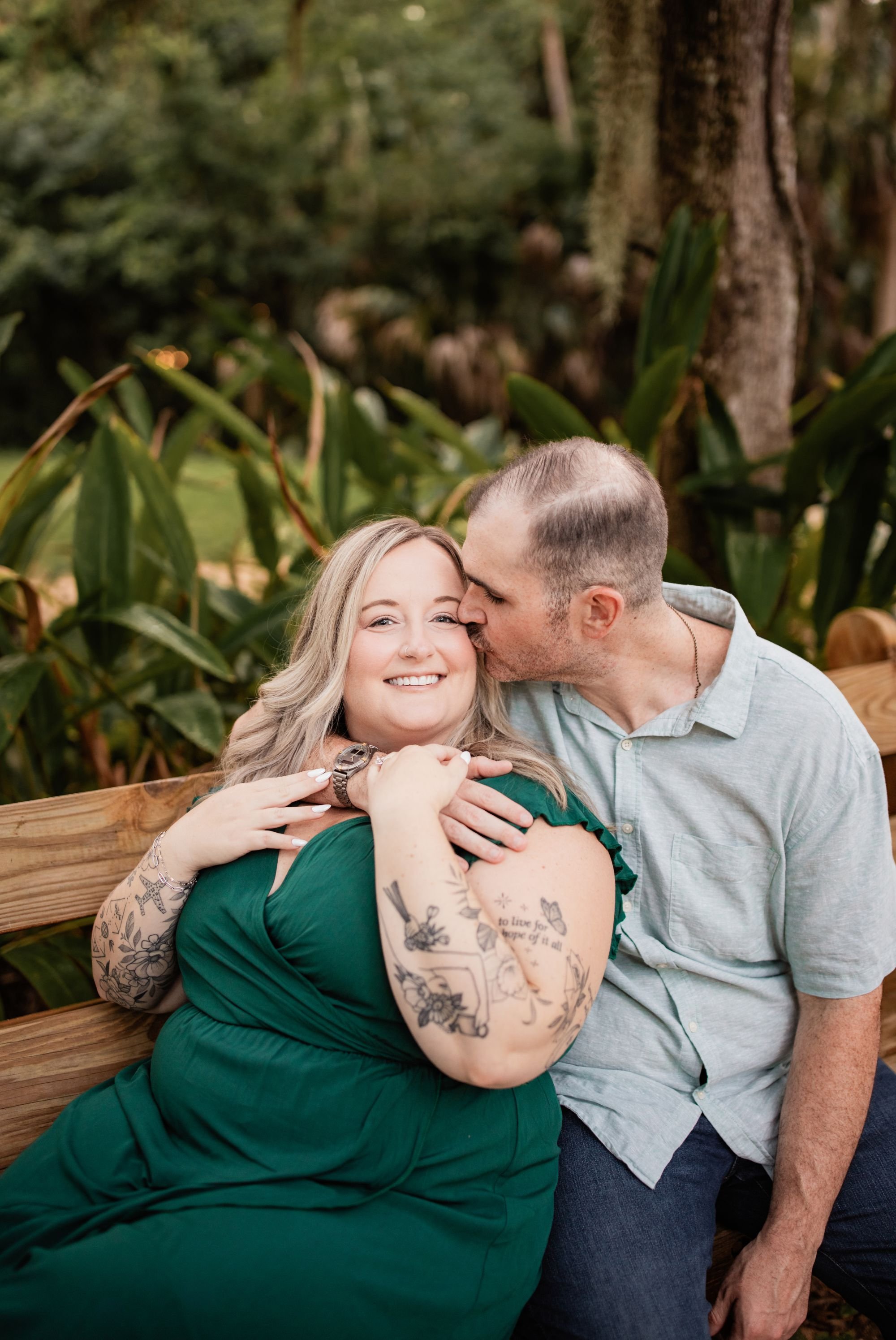Bride-to-be smiling as her fiancé kisses her cheek while they sit together on a garden bench.