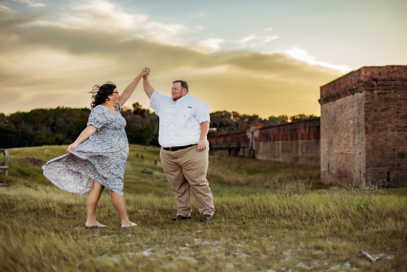 Couple dancing in a grassy field in a patterned dress and neutral shirt near a historic fort