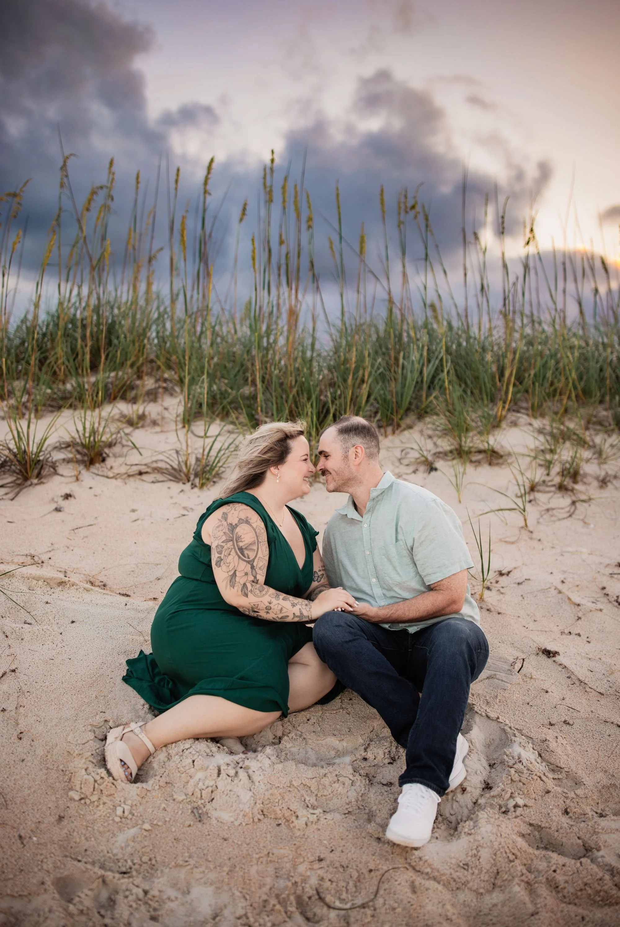 Engaged couple sitting together in the sand dunes at Washington Oaks beach beneath a cloudy Florida sky.