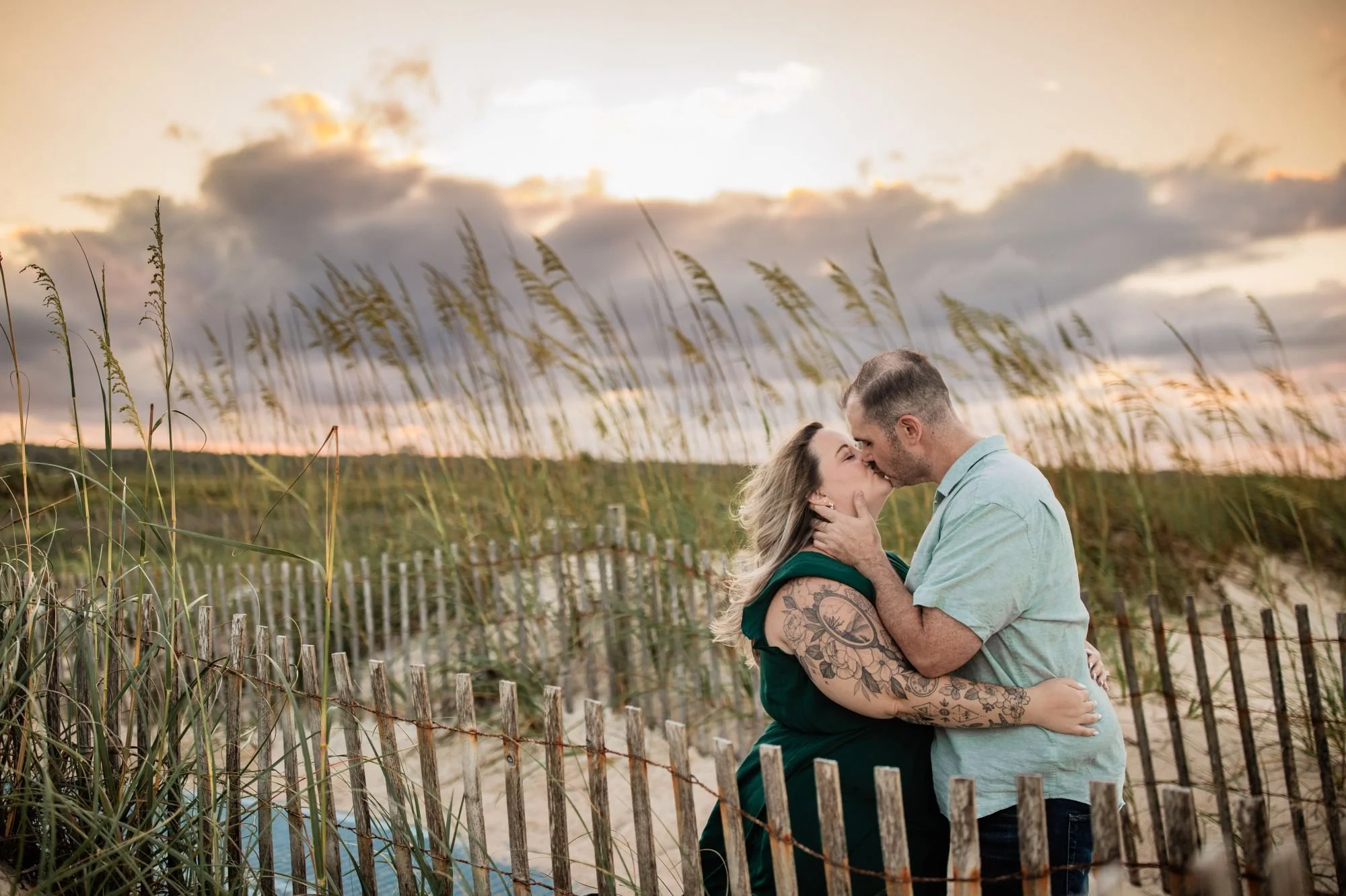 Engaged couple kissing beside the dune fence and blowing sea oats under a dramatic sunset sky at Washington Oaks beach.
