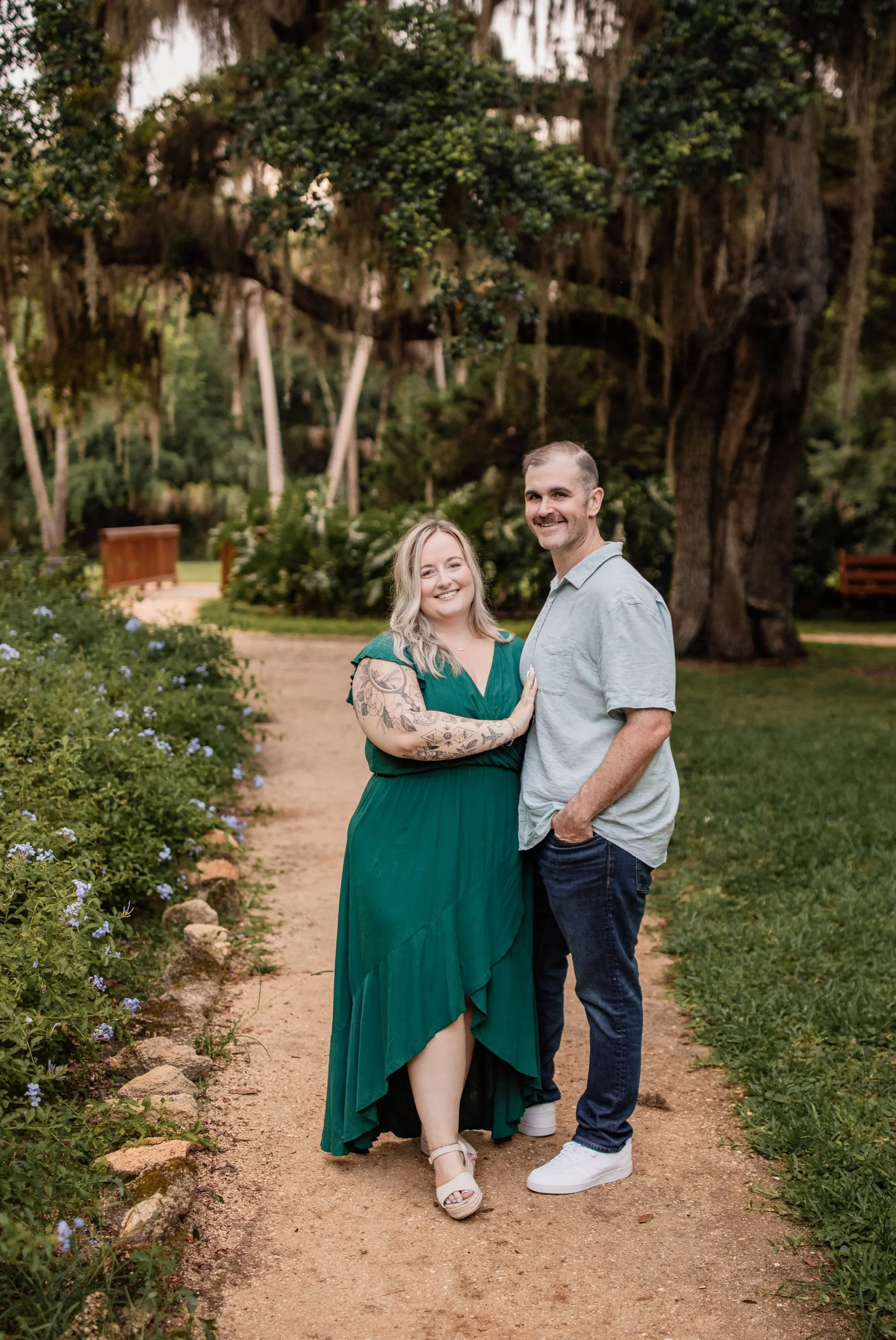 Engaged couple smiling together on a garden path surrounded by flowers and large oak trees.