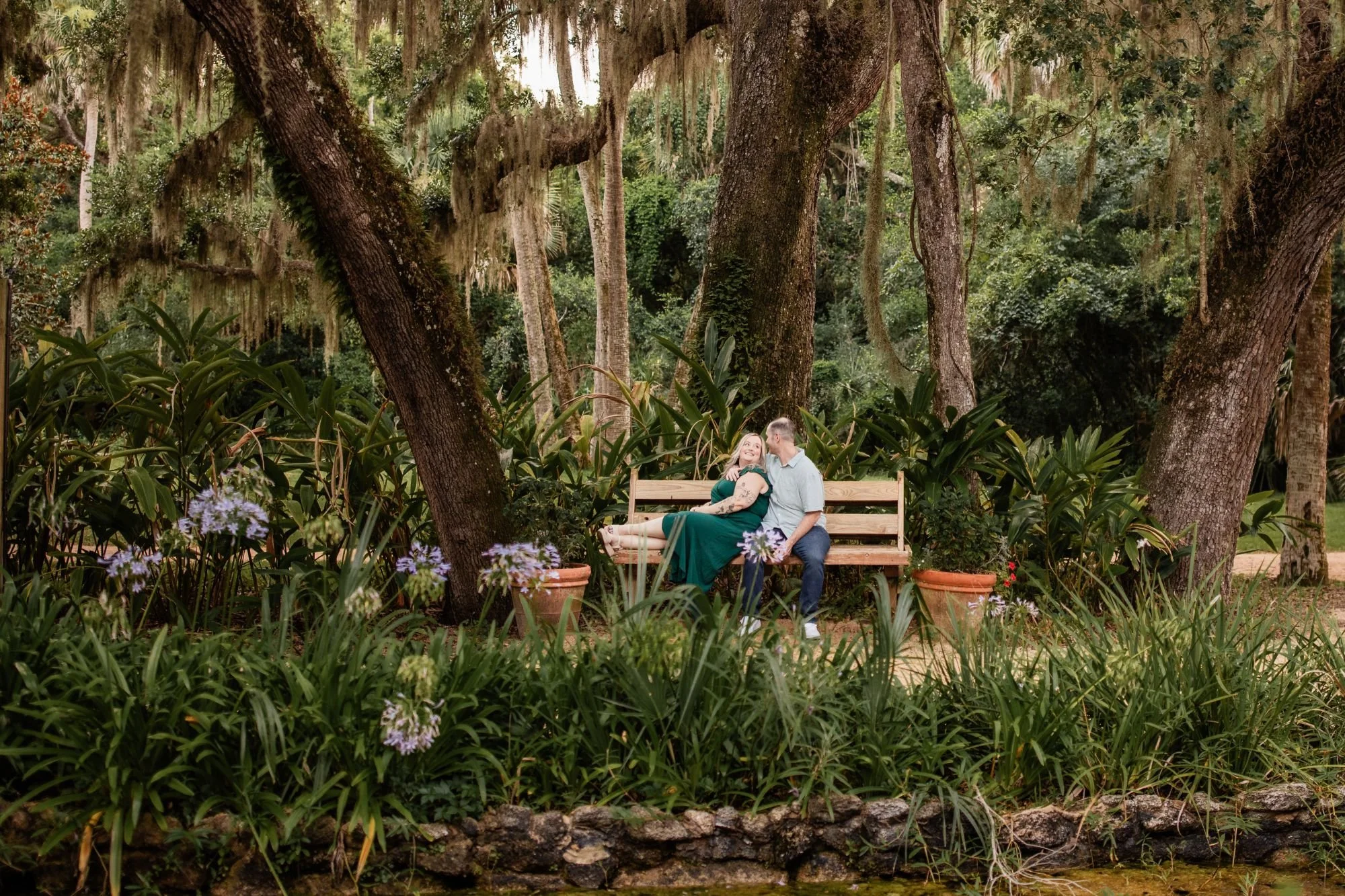Engaged couple laughing together on a bench surrounded by oak trees, flowers, and lush gardens.