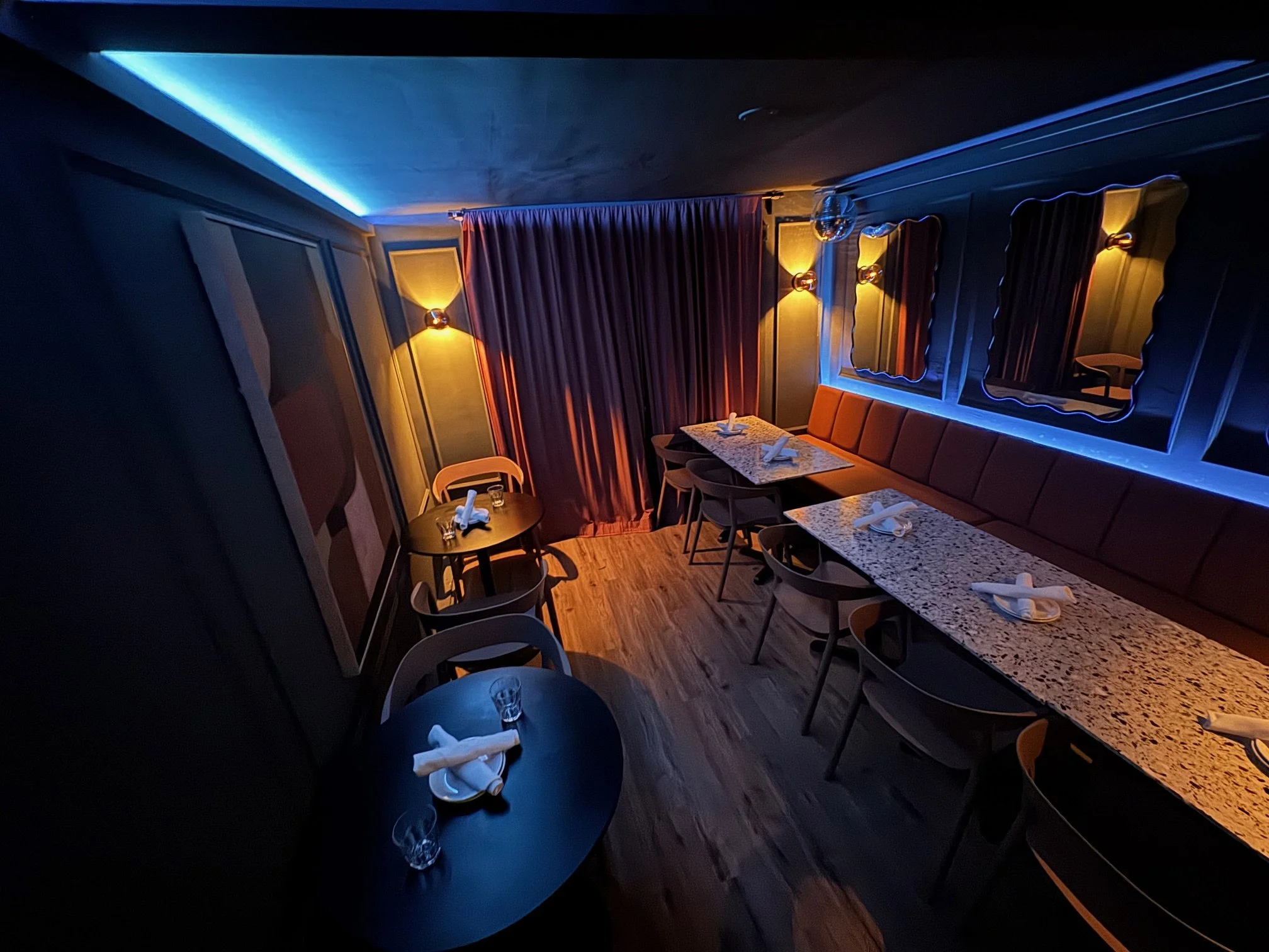 Dimly lit restaurant with a long counter, small tables set with napkins and glasses, wall mirrors, wall sconces, and curtains creating a cozy atmosphere.