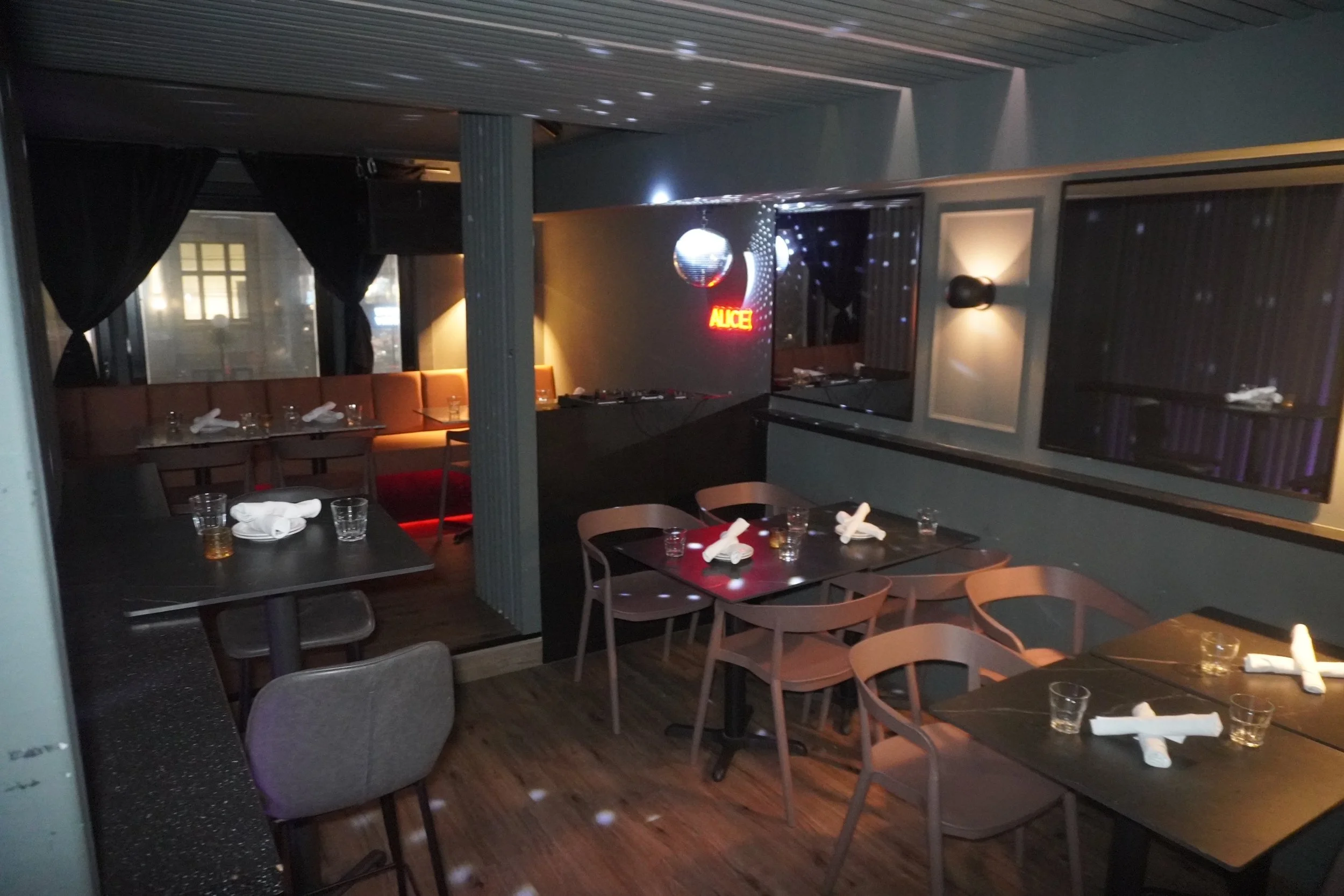 Restaurant with tables set for dining, napkins rolled on plates, glasses, and a disco ball with a sign that reads "ALICE" on the wall.