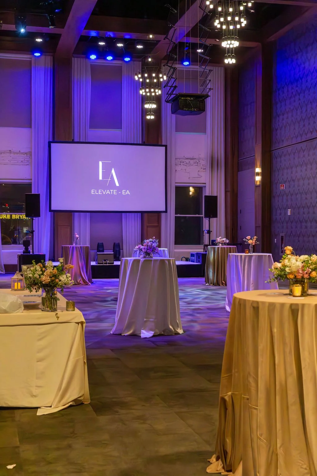 A decorated event hall with high ceilings, purple lighting, floral arrangements on round tables, and a large screen displaying the logo "ELEVATE - EA."