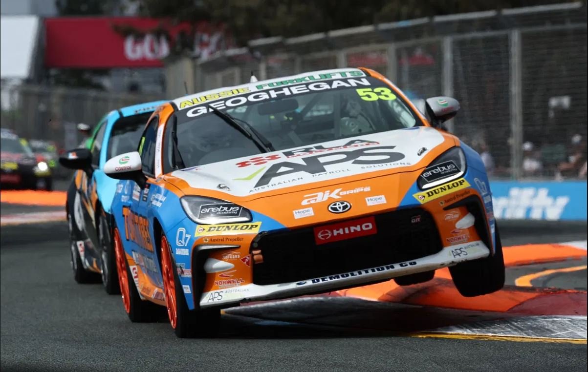 A race car flying over a track curb during a race, with a blurred background of other racing cars and fences.