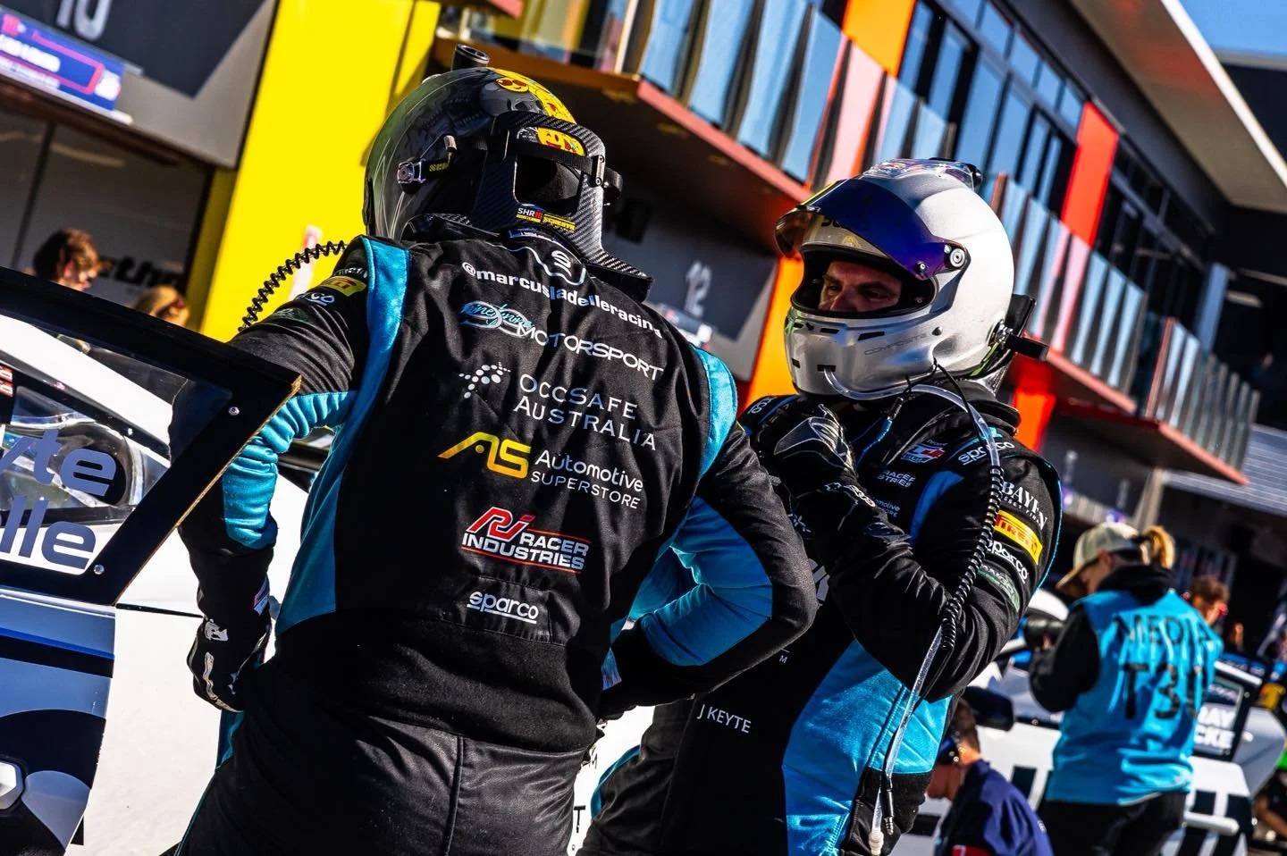 Two race car drivers in racing suits and helmets talking beside a race car, with pit crew members and a modern building in the background.
