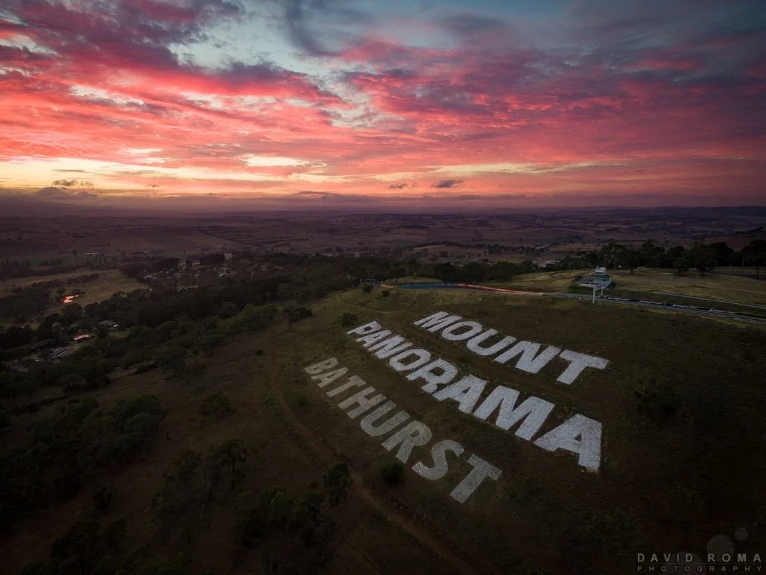 Sunset over a hillside with large white text reading 'MOUNT PANORAM A BAT HURST' on the slope.