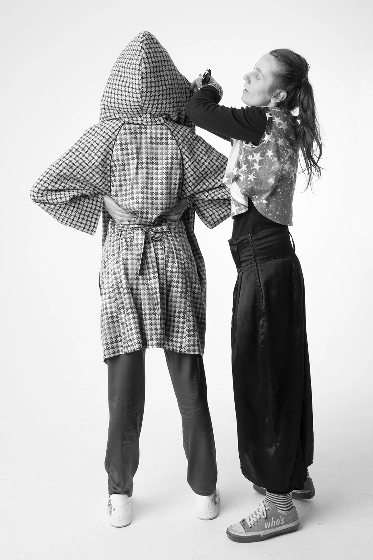 Black and white photo of two women standing close, one with a hooded patterned coat and the other with a star-patterned jacket, holding a camera or phone, with one woman wearing sneakers and the other in shoes with striped socks.