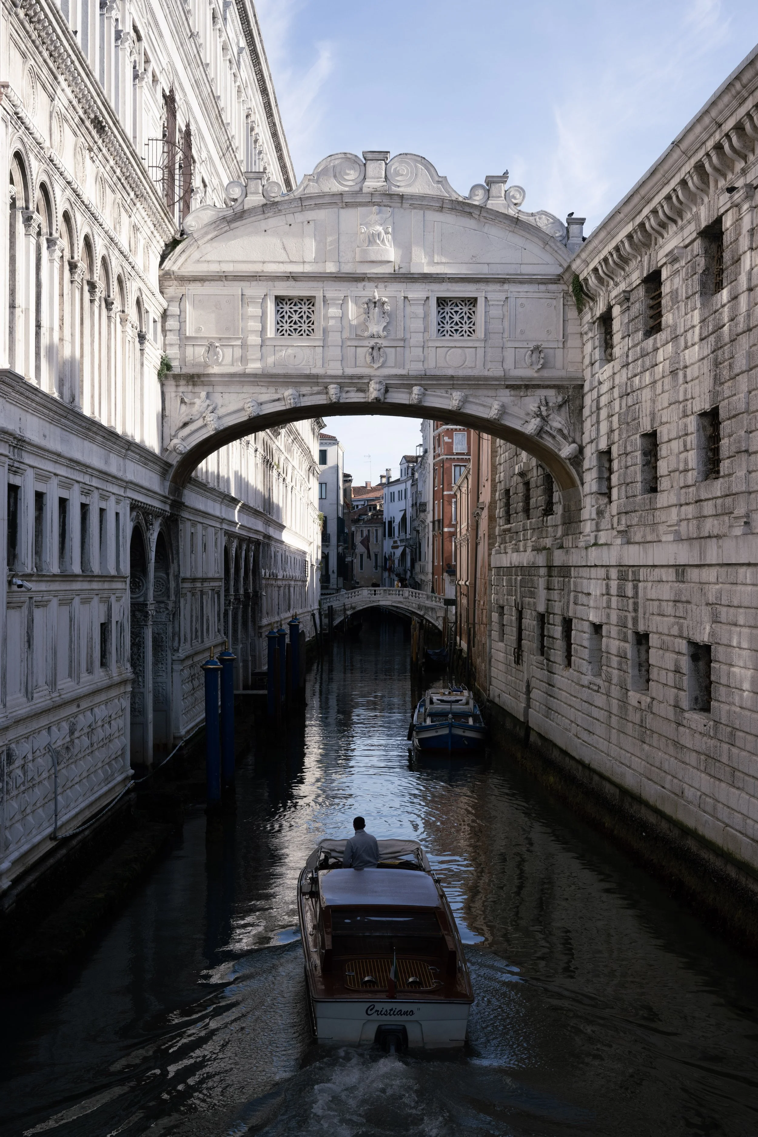A canal with a boat and a person steering, surrounded by historic buildings, with the Bridge of Sighs crossing overhead in Venice, Italy.